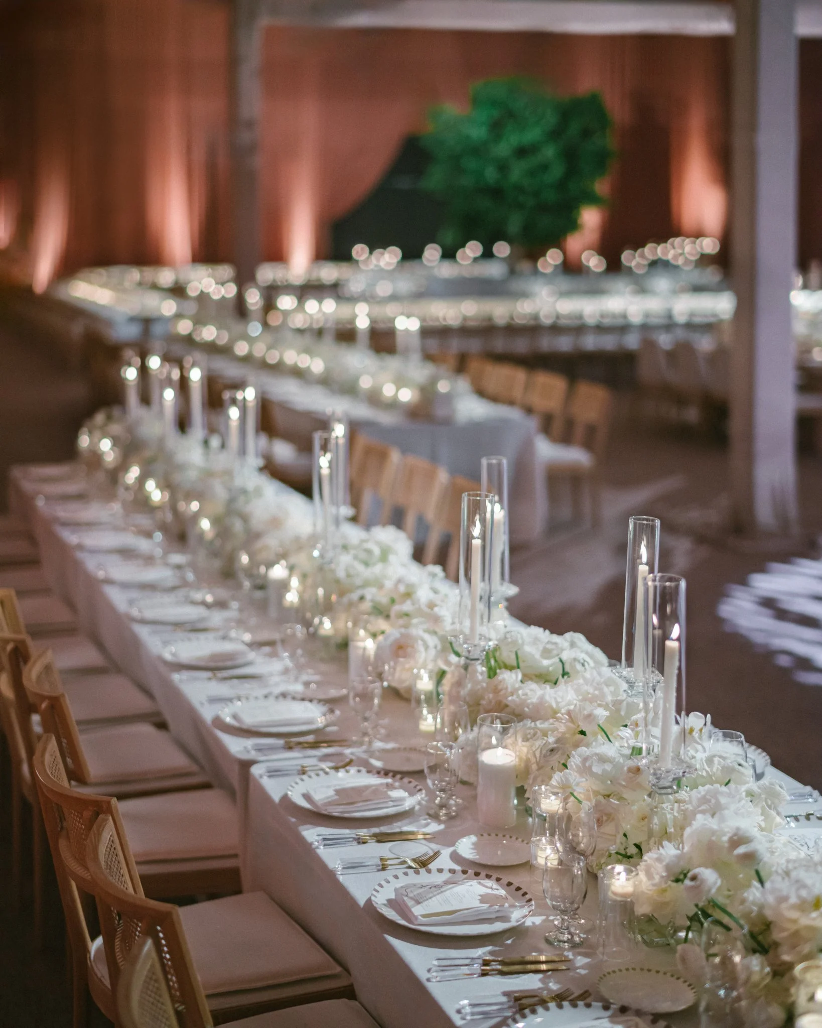Elegant wedding reception table decorated with white flowers, candles, glassware, and gold accents in a rustic barn setting with warm lighting.