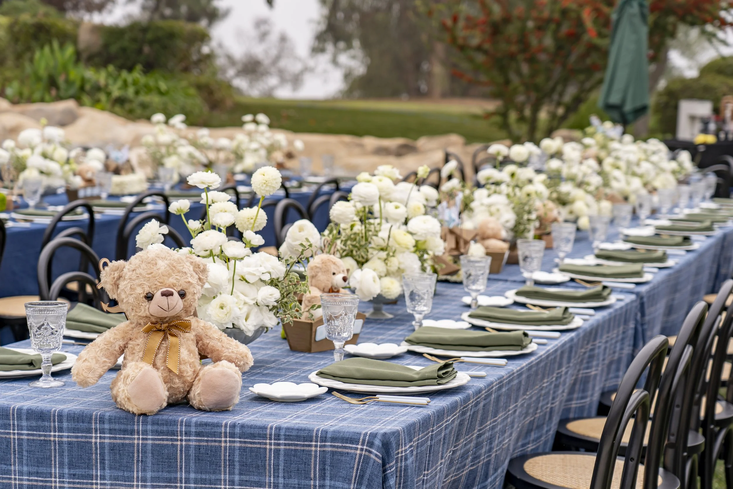 Long outdoor dining table decorated with white flowers, teddy bears, glassware, and green napkins, set for a celebration in a garden area.