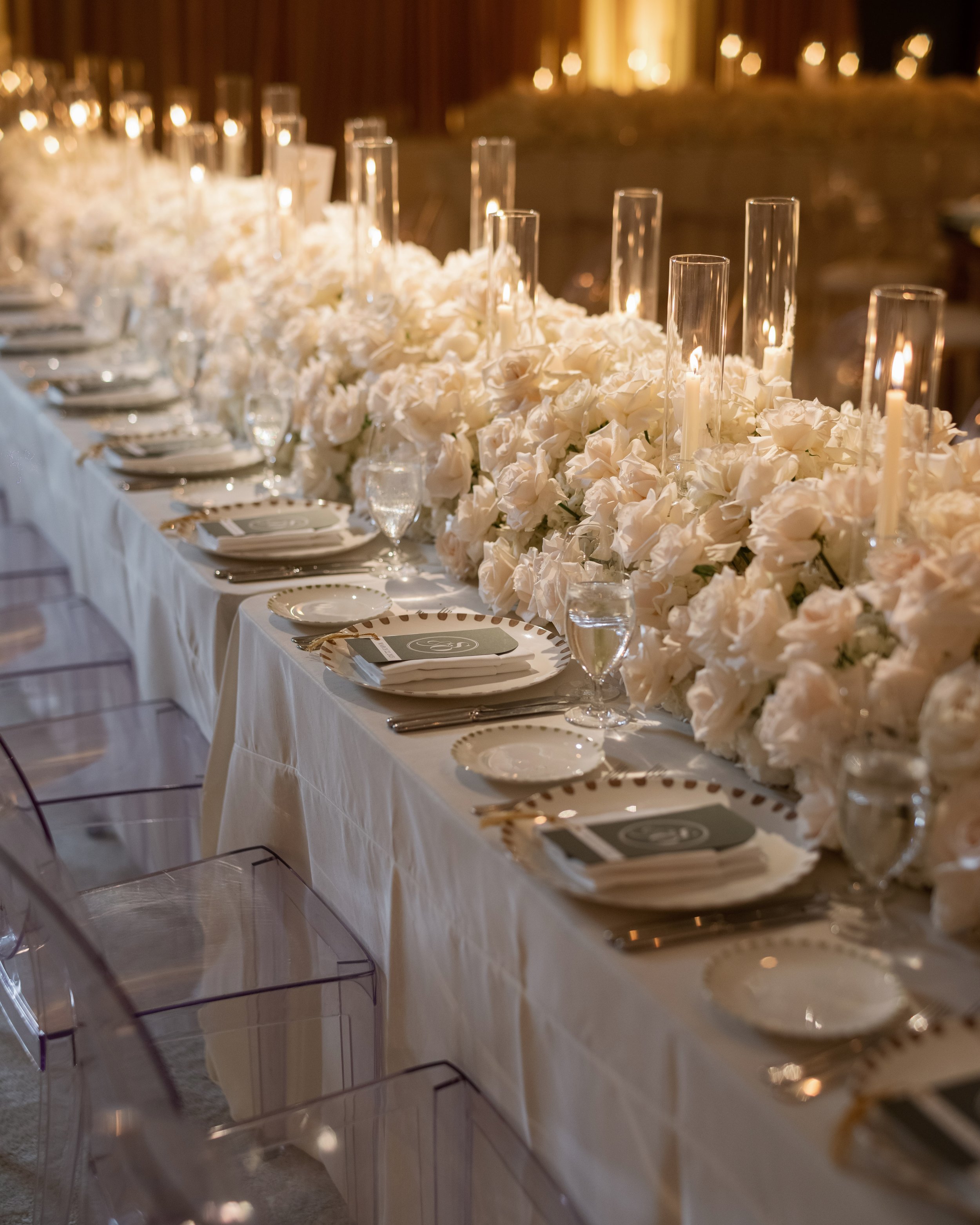 Elegant wedding reception table decorated with a long white tablecloth, white floral arrangements, glass candles, and place settings with plates, silverware, and water glasses.