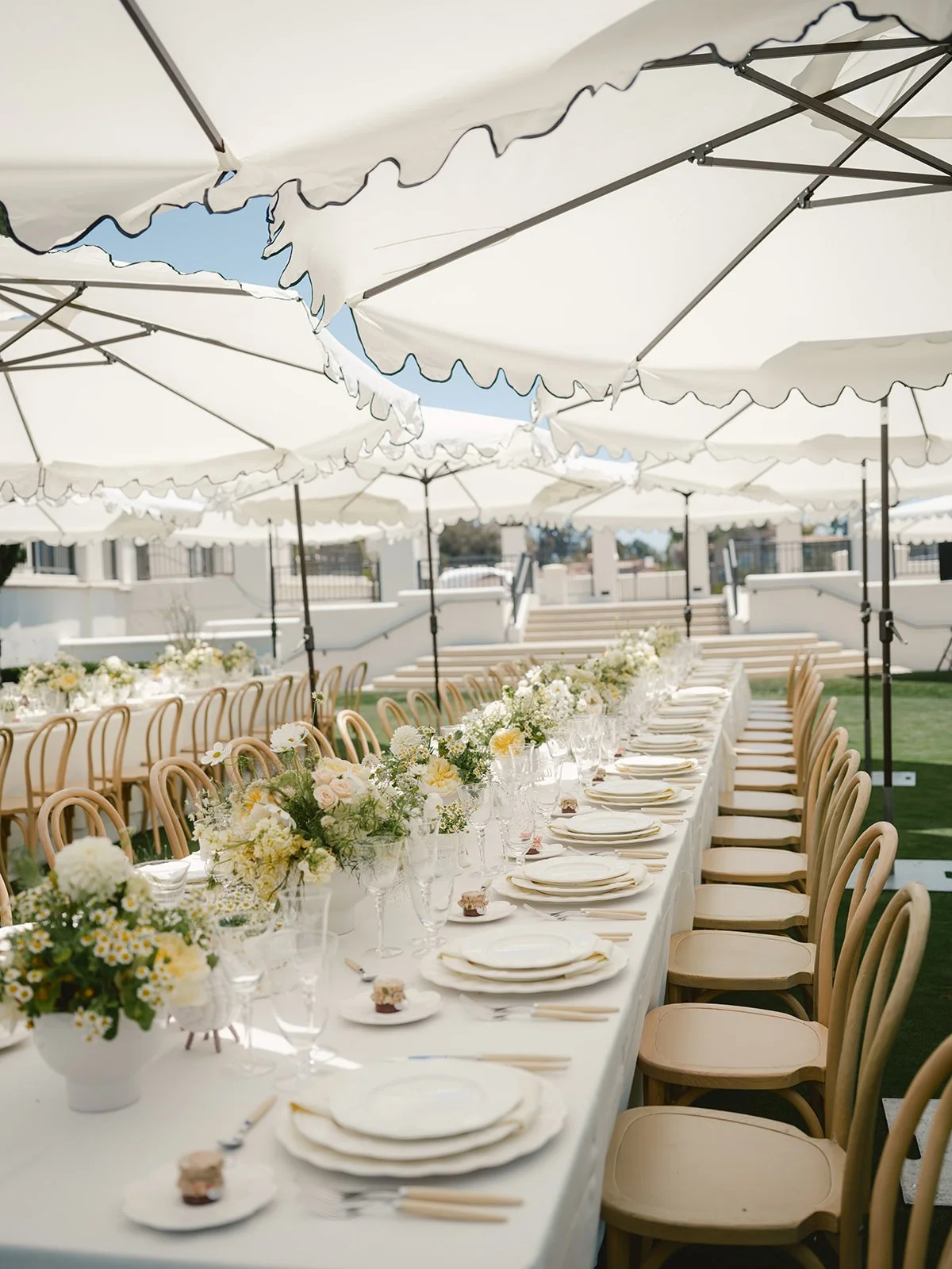 Long outdoor dining table under white umbrellas, decorated with bouquets of white, yellow, and pink flowers, set with plates, glasses, and utensils, in a sunny outdoor setting.