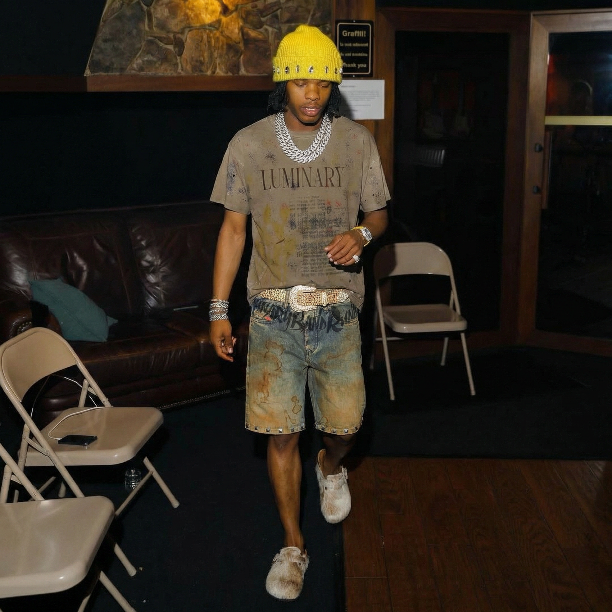 A young man wearing a yellow beanie, a distressed gray T-shirt with the word 'LUMINARY', and multicolored denim shorts, walking past beige chairs in a dimly lit room with wooden walls and a dark floor.