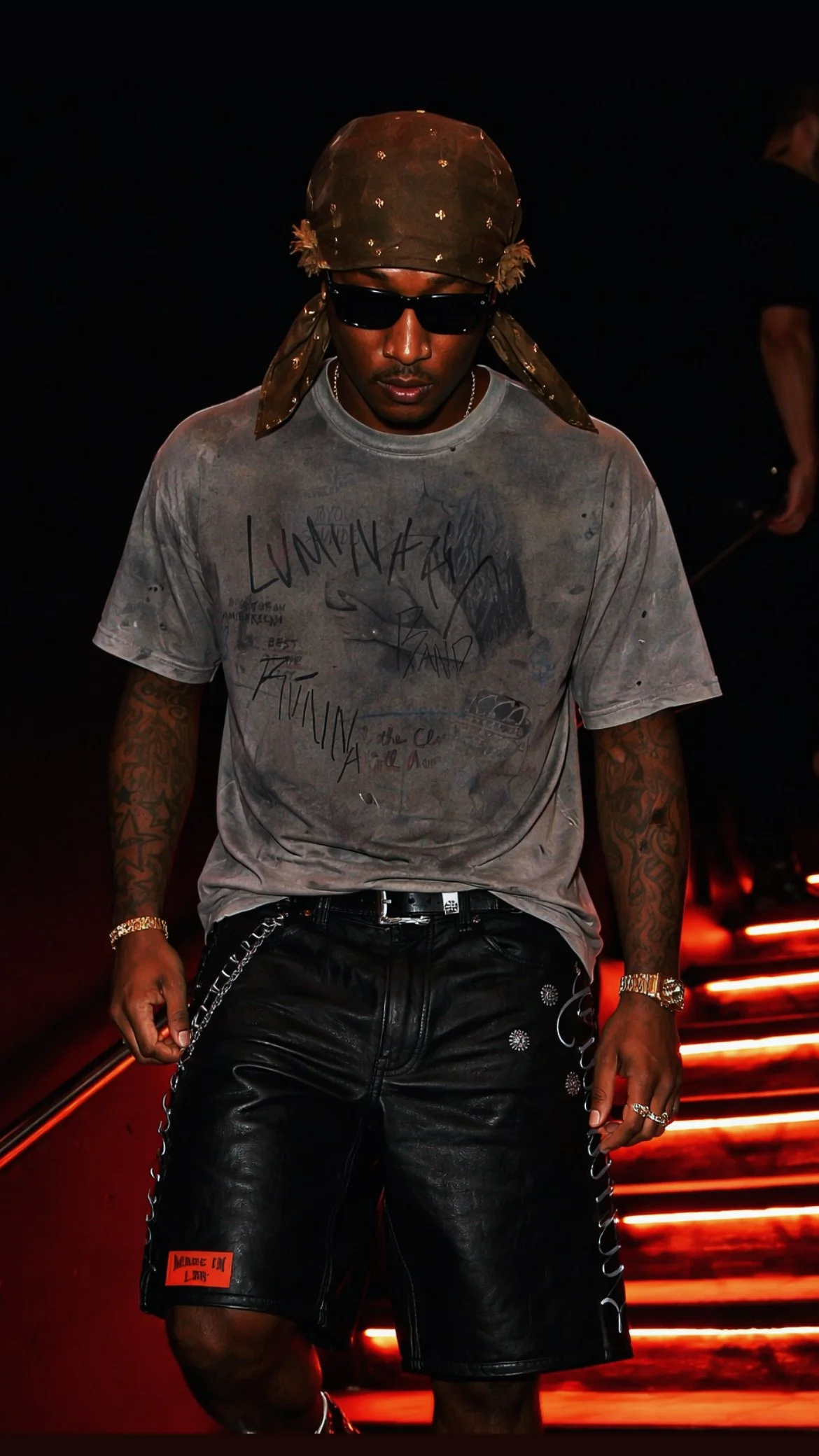 A man wearing sunglasses, a brown bandana, a distressed graphic t-shirt, black leather shorts with chains, and jewelry, standing on illuminated stairs in a dark setting.