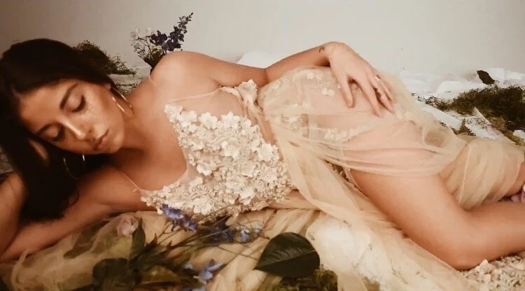 A woman lying on a bed with her eyes closed, wearing a sheer, floral-embellished dress, with flowers and plants around her.