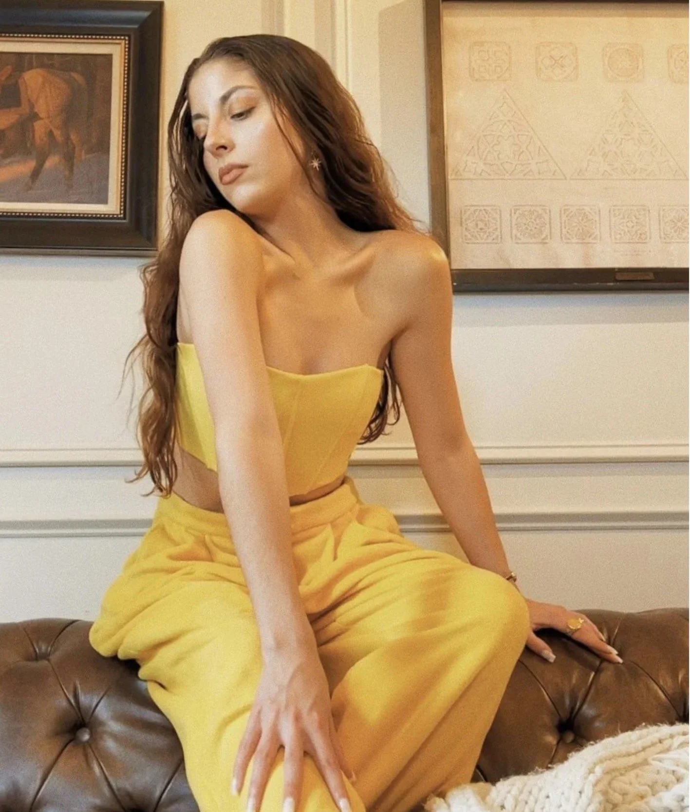 A young woman with long brown hair, wearing a yellow strapless top and matching yellow pants, sitting on a brown leather couch in an indoor setting with framed artwork on the walls.