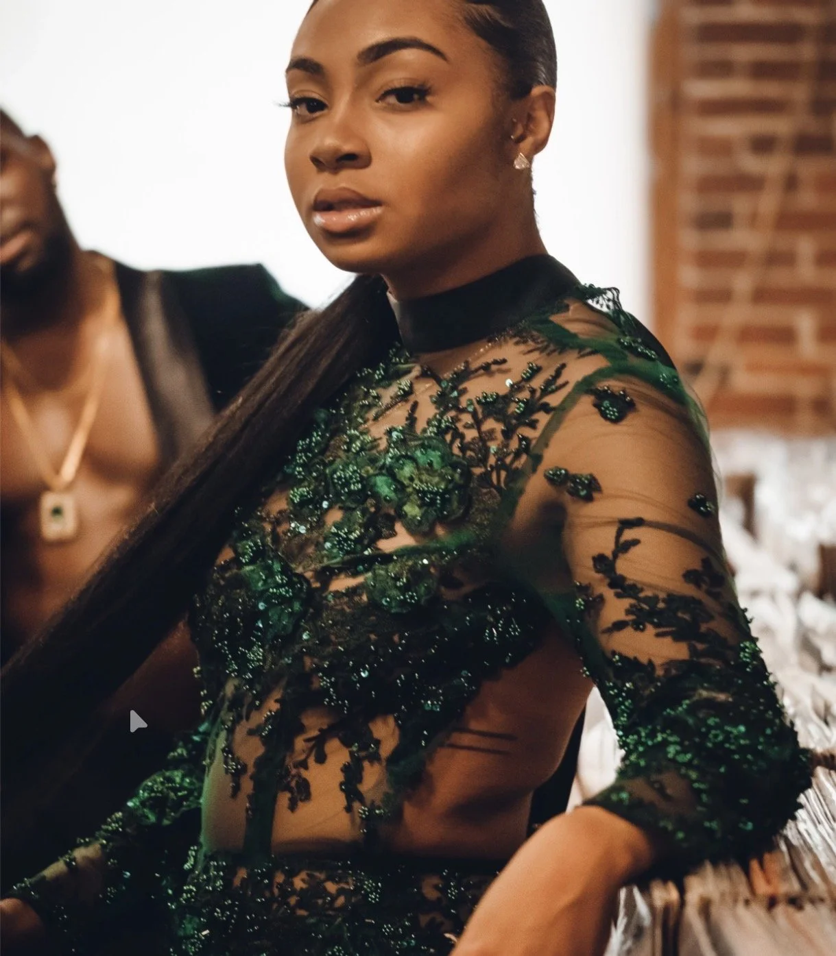 A woman wearing a sheer dress with green beaded embroidery poses indoors, with a man with a beard and gold chain in the background.