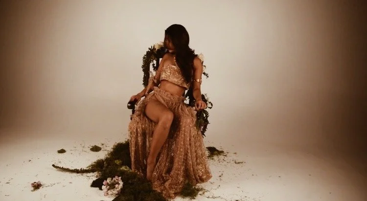 Woman sitting on a throne-like seat decorated with greenery and flowers, wearing a gold outfit, in a studio with a plain background.