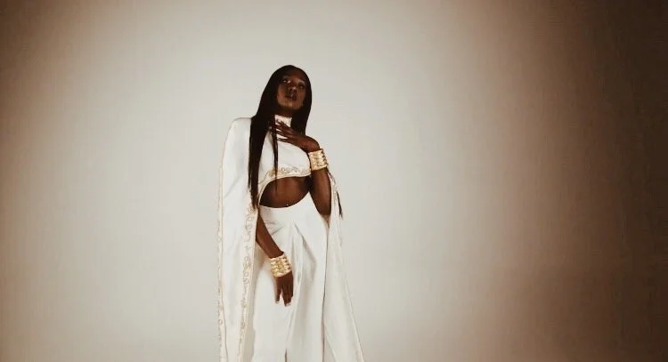 A woman in white attire with long dark hair, standing against a plain background, holding her hand to her chest, wearing multiple gold bangles.