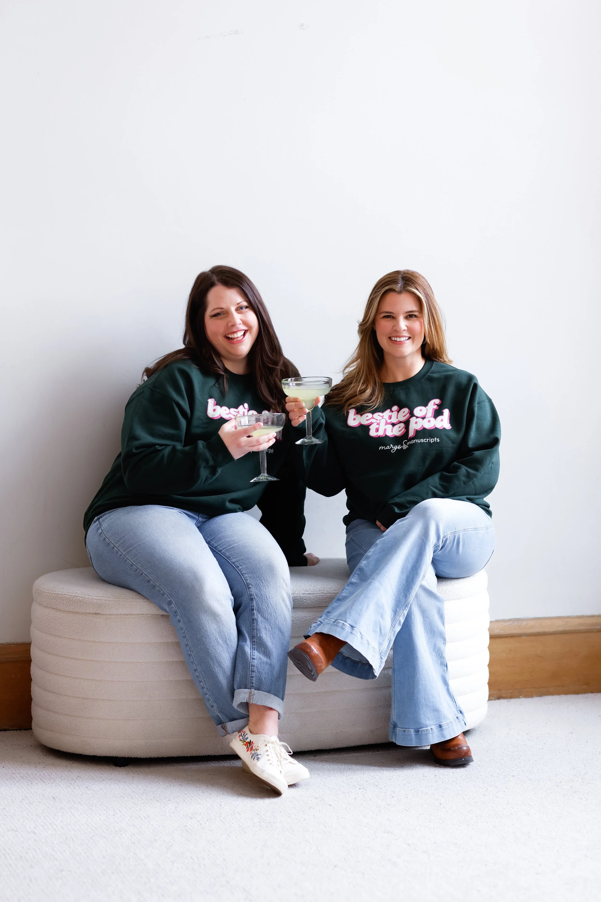 Two women sitting on a round, white, textured ottoman, smiling and holding cocktails, against a plain white wall. Both are wearing matching dark green sweatshirts with pink and white text, light blue jeans, and casual shoes.
