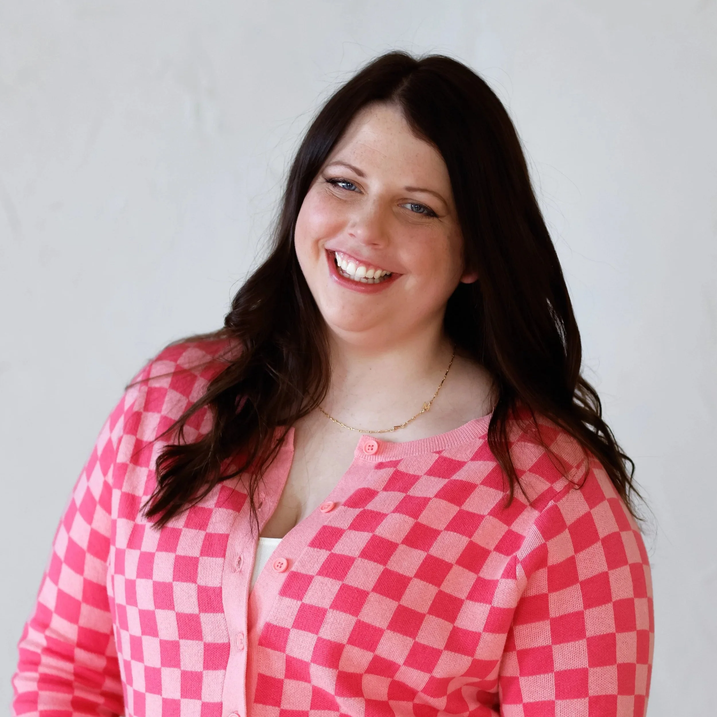 A woman with long dark hair wearing a pink checkered cardigan, smiling, standing against a plain gray background.
