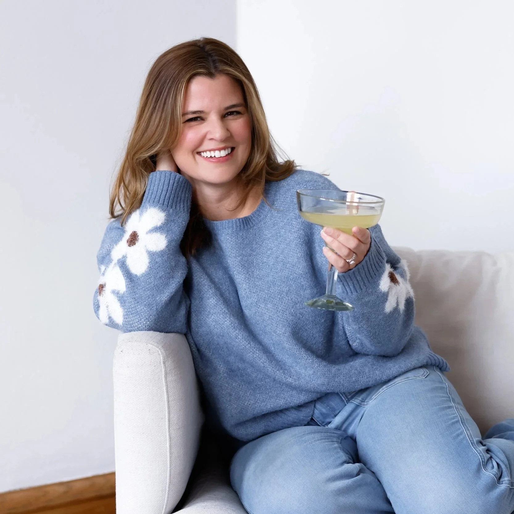 A woman sitting on a white sofa, smiling, wearing a blue sweater with white floral patterns on the sleeves, holding a cocktail glass with a light-colored drink in an indoor setting.
