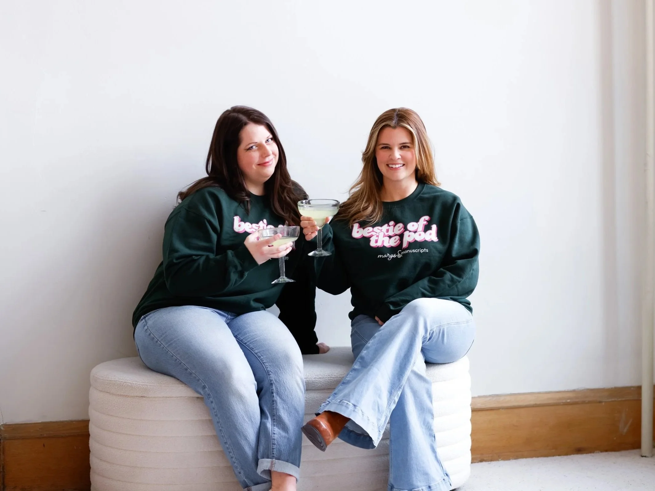 Two women sitting on a white ottoman, wearing matching green sweatshirts that say 'bestie of the pod,' holding champagne glasses, smiling at the camera, in a minimalistic room with a plain white wall and wooden baseboard.