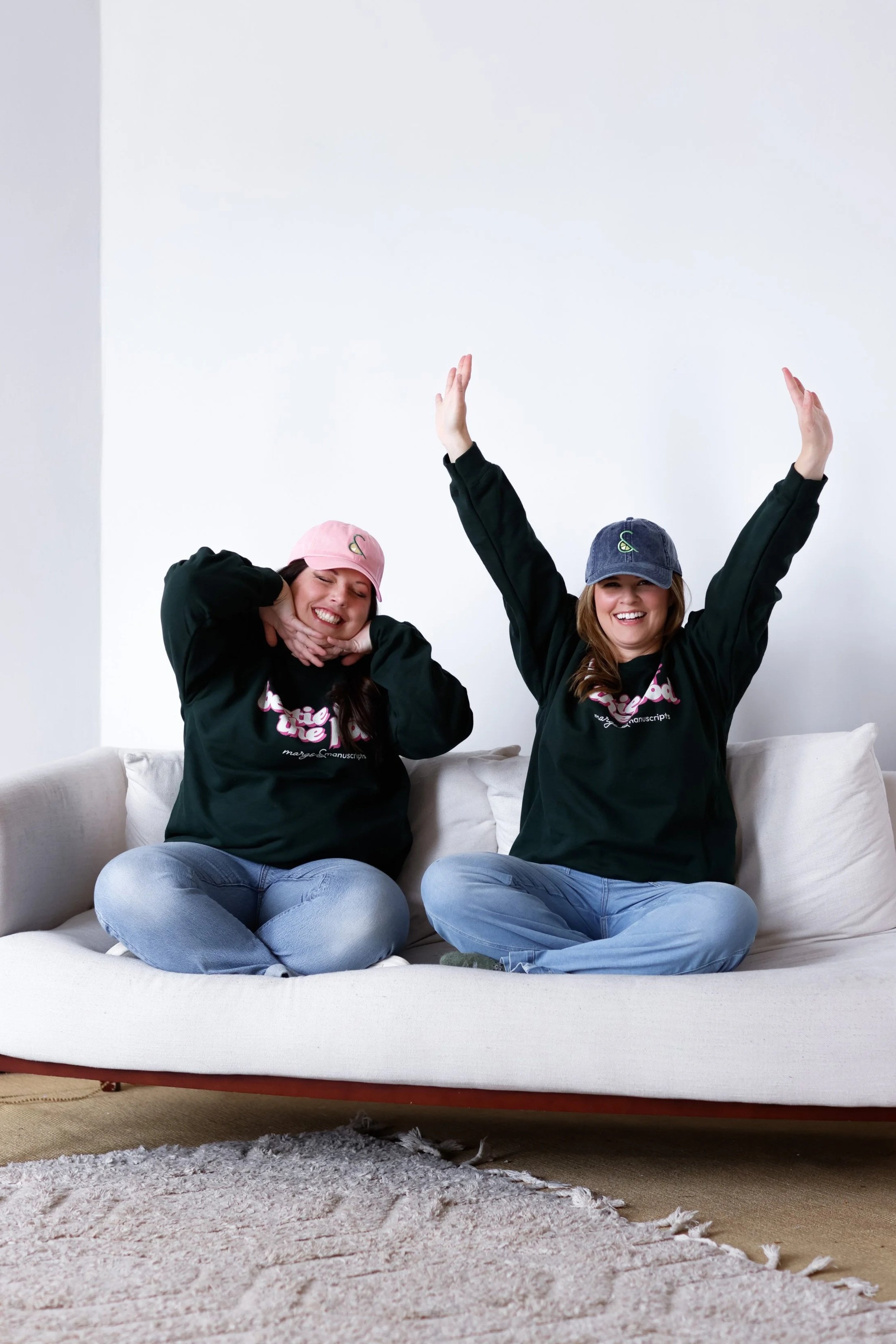 Two women sitting on a white sofa, smiling and posing with their arms raised and crossed. They are wearing black sweatshirts and baseball caps, one pink and one blue, in a bright room with a white wall background.