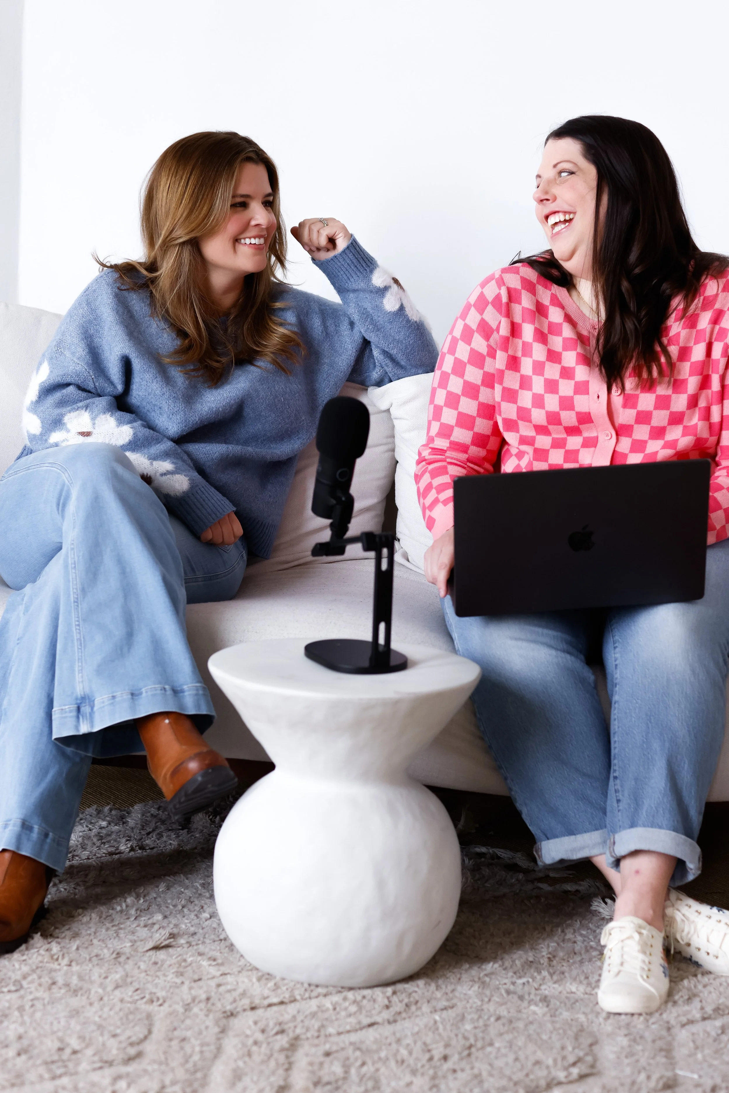 Two women sitting on a couch, one making a muscle, with a microphone on a small table between them, and a laptop on the lap of one woman, in a cozy living room.