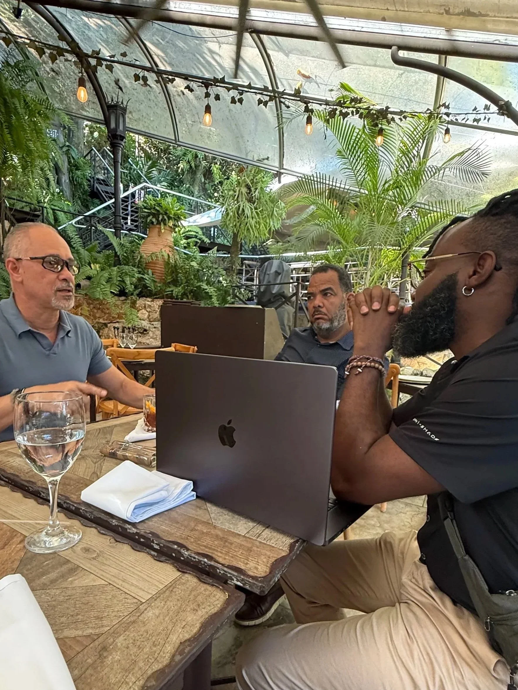 Three men sitting at a wooden table having a discussion in a greenhouse-like setting with lush green plants and string lights overhead, a laptop open on the table.