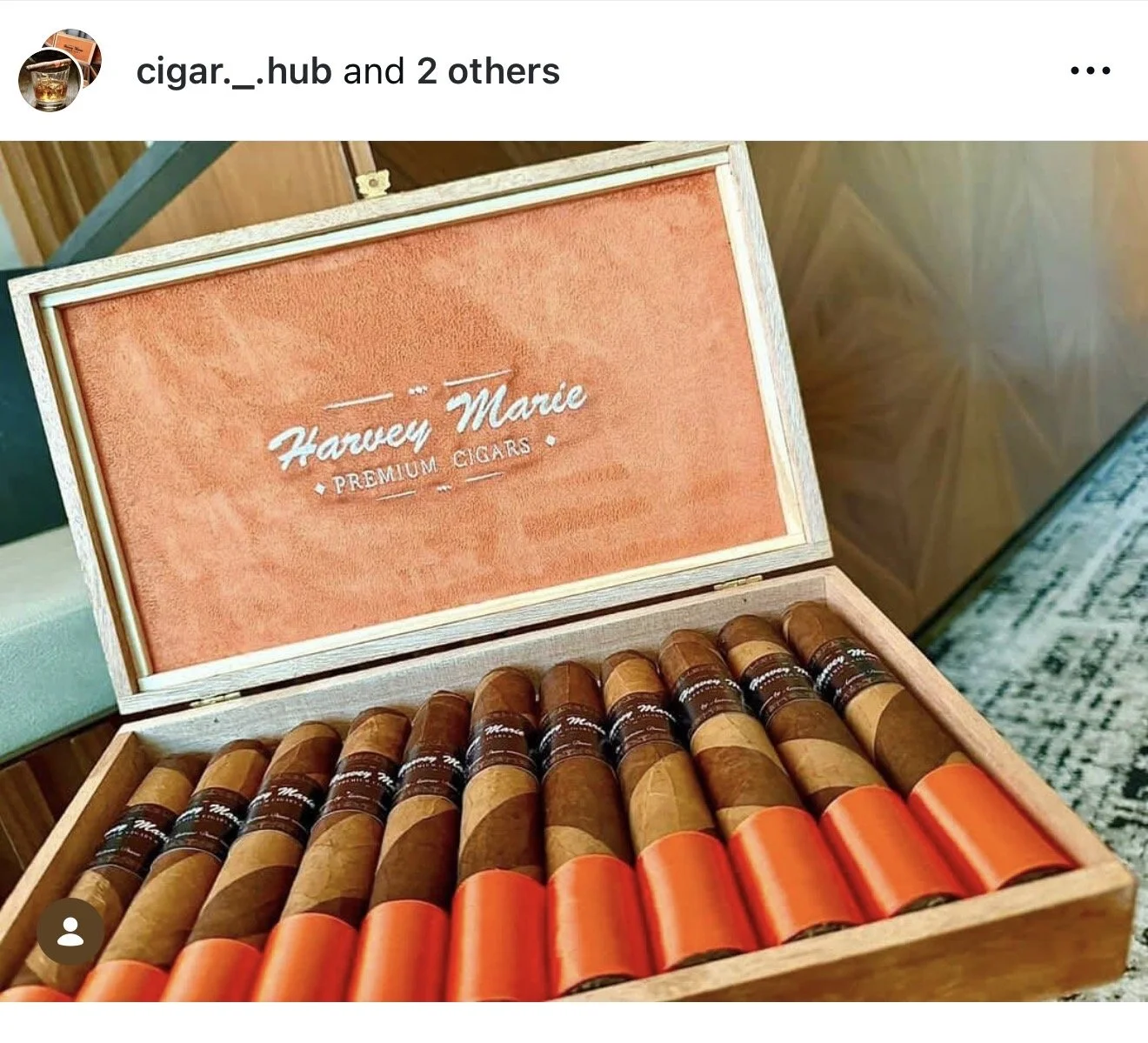 Open wooden box of cigars labeled "Harvey Marle Premium Cigars" with a pink interior lining. Inside are cigars with orange and brown bands.