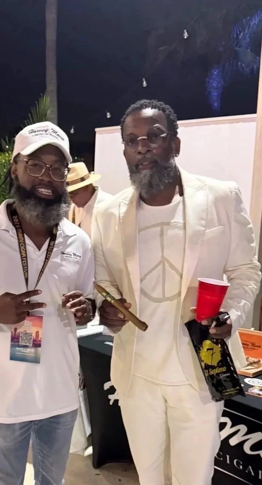 Two men with gray beards and glasses posing together at an event, one wearing a white baseball cap and the other in a cream suit, holding a cigar and a red cup, with a woman in a hat in the background.