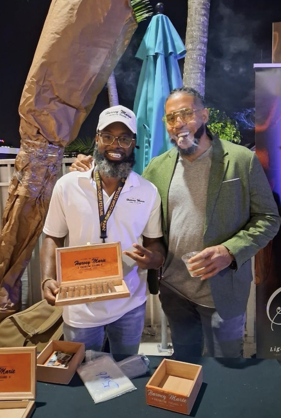 Two men smiling, one holding a box of cigars, the other holding a drink, at an outdoor event with decorative elements and a colorful umbrella in the background.