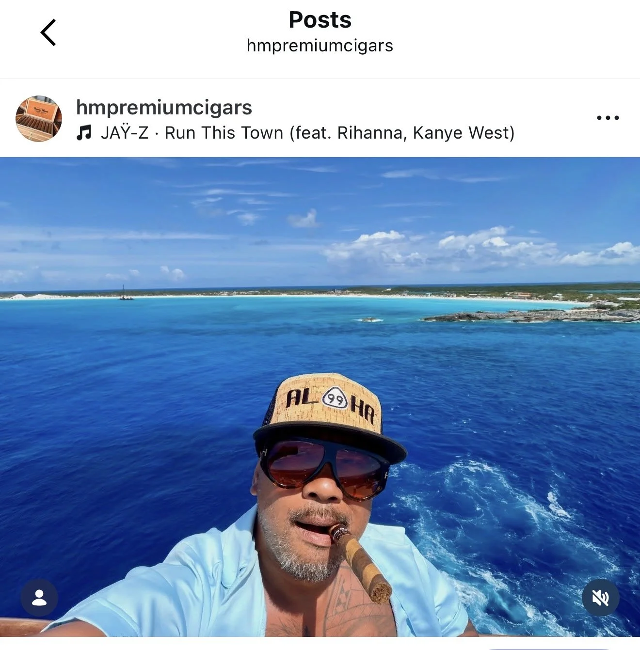 A man wearing sunglasses and a tan cap with text, holding a cigar in his mouth, taking a selfie on a boat with a large body of blue water and a distant shoreline with white sandy beaches and a partly cloudy sky in the background.