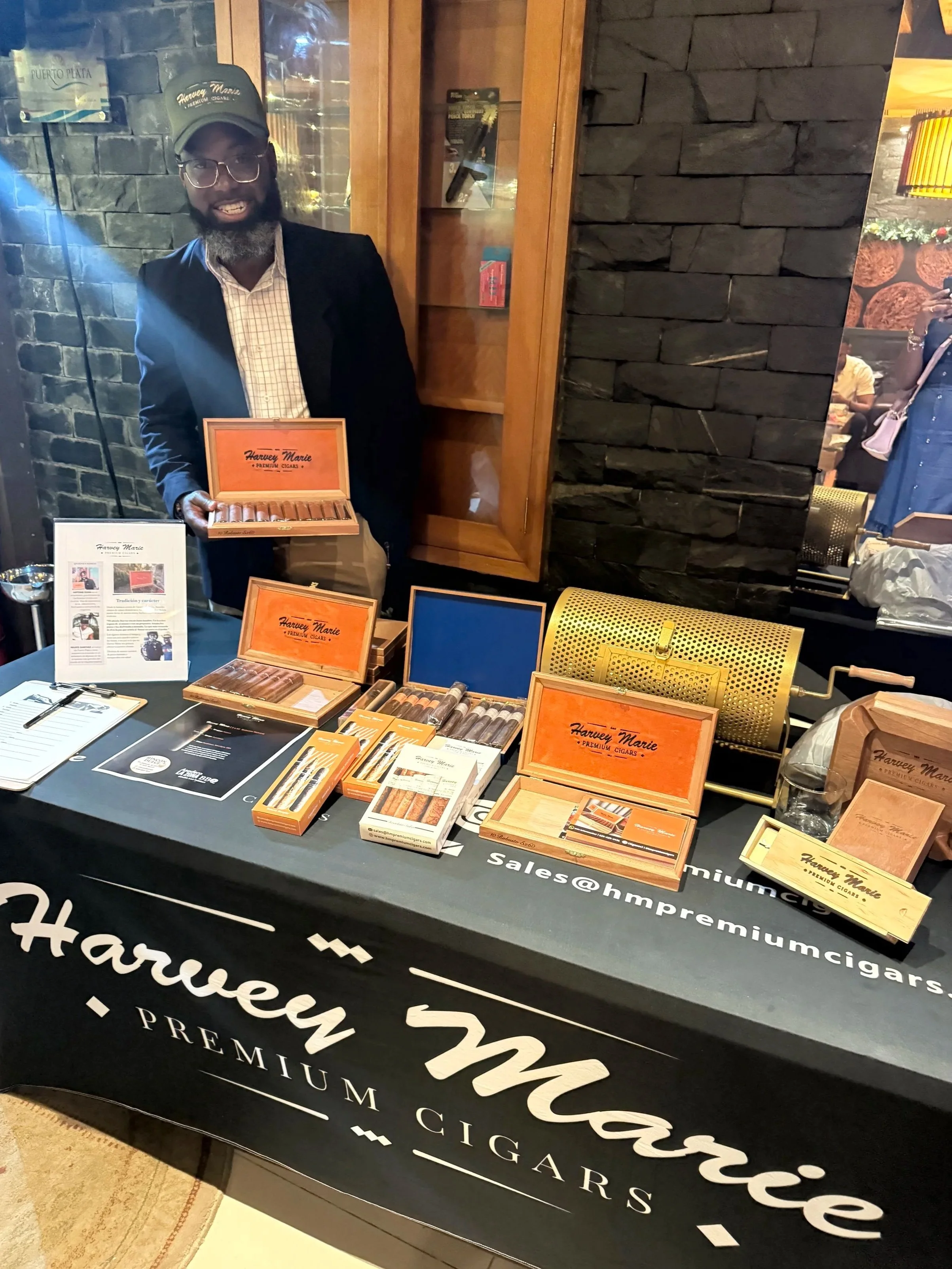 A man with glasses and a beard, wearing a black cap and blazer, stands behind a table displaying Harvey Marie premium cigars. The table has cigar boxes, promotional materials, and a black cloth with the Harvey Marie logo and contact information.