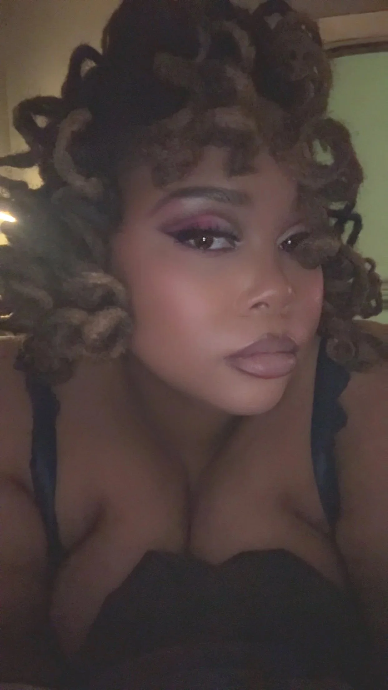 A woman with curly, highlighted hair and makeup, wearing a black top with thin straps.