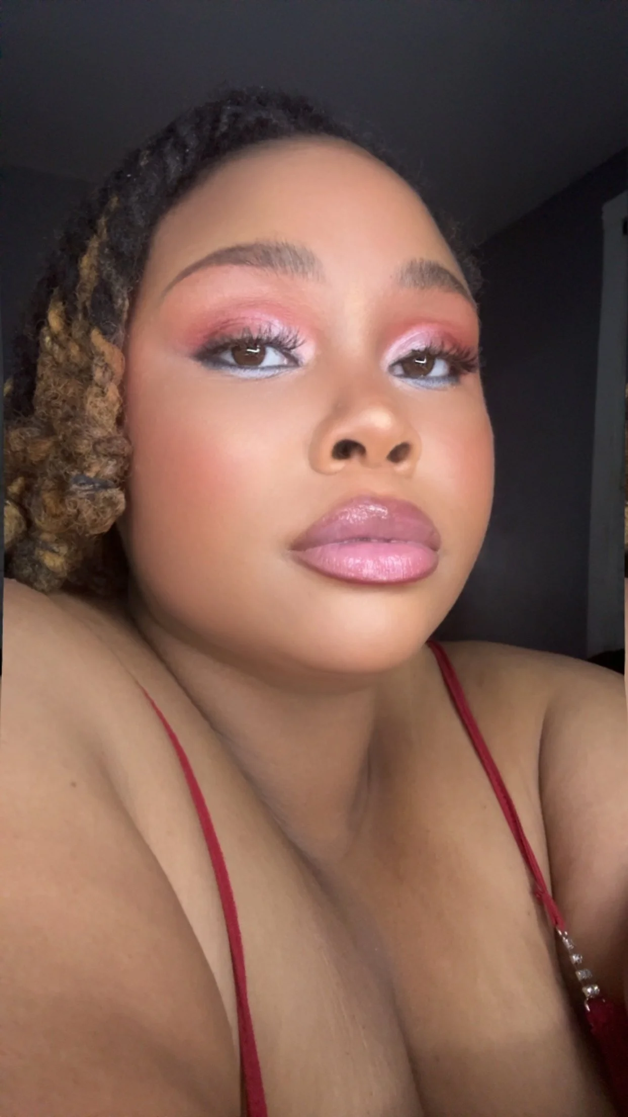 Close-up of a woman with makeup, wearing a red spaghetti strap top, looking at the camera.
