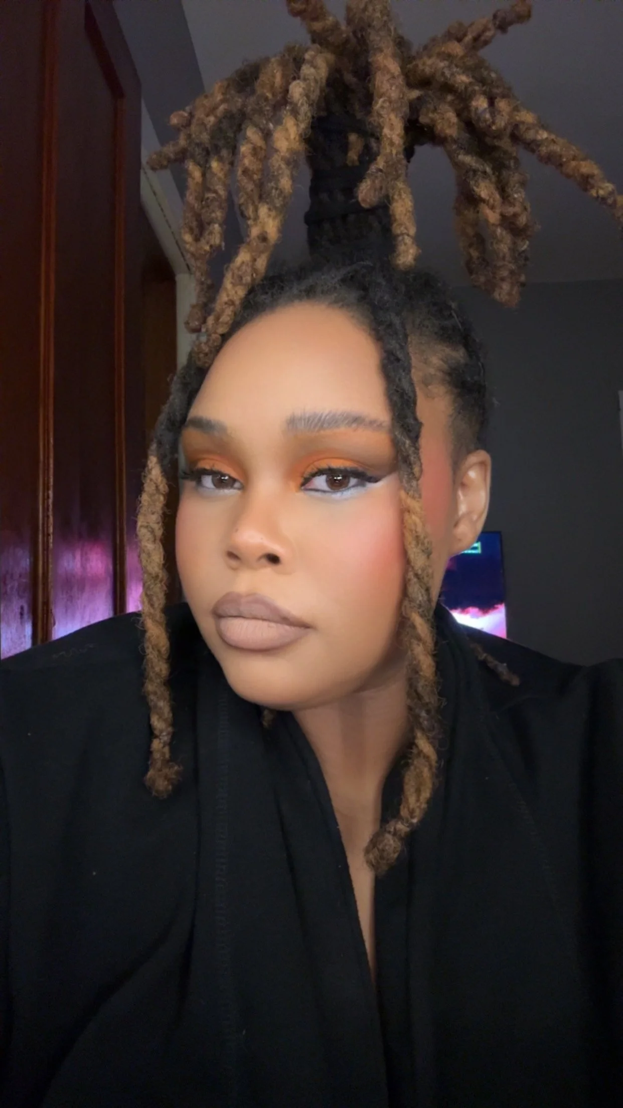 A woman with styled dreadlocks updo, wearing makeup with bright eyeshadow, lipstick, and blush, looking into the camera, indoors with a background of wooden cabinets and a television.