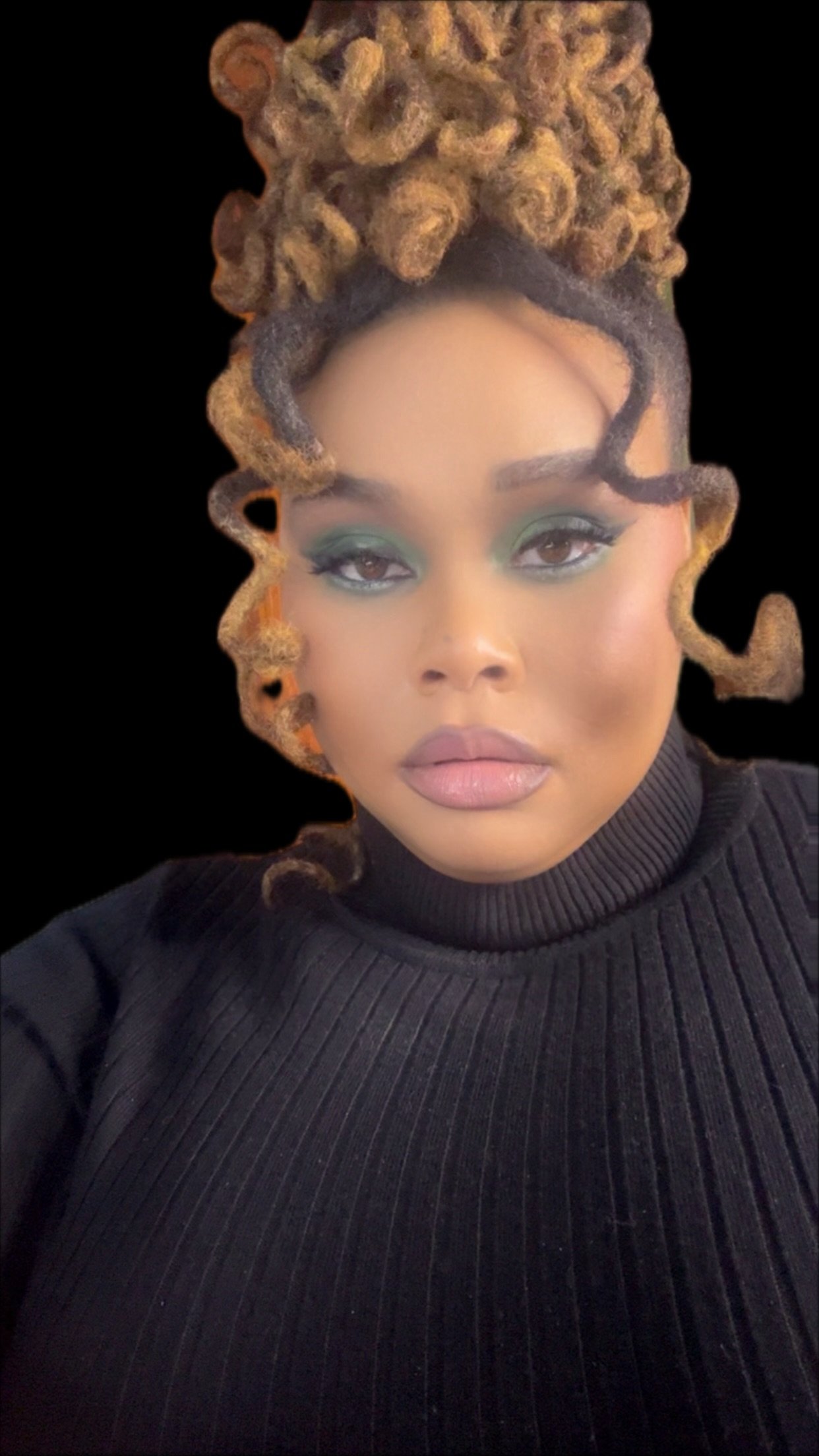 A woman with styled, curly dreadlocks and makeup, wearing a black turtleneck top, against a black background.