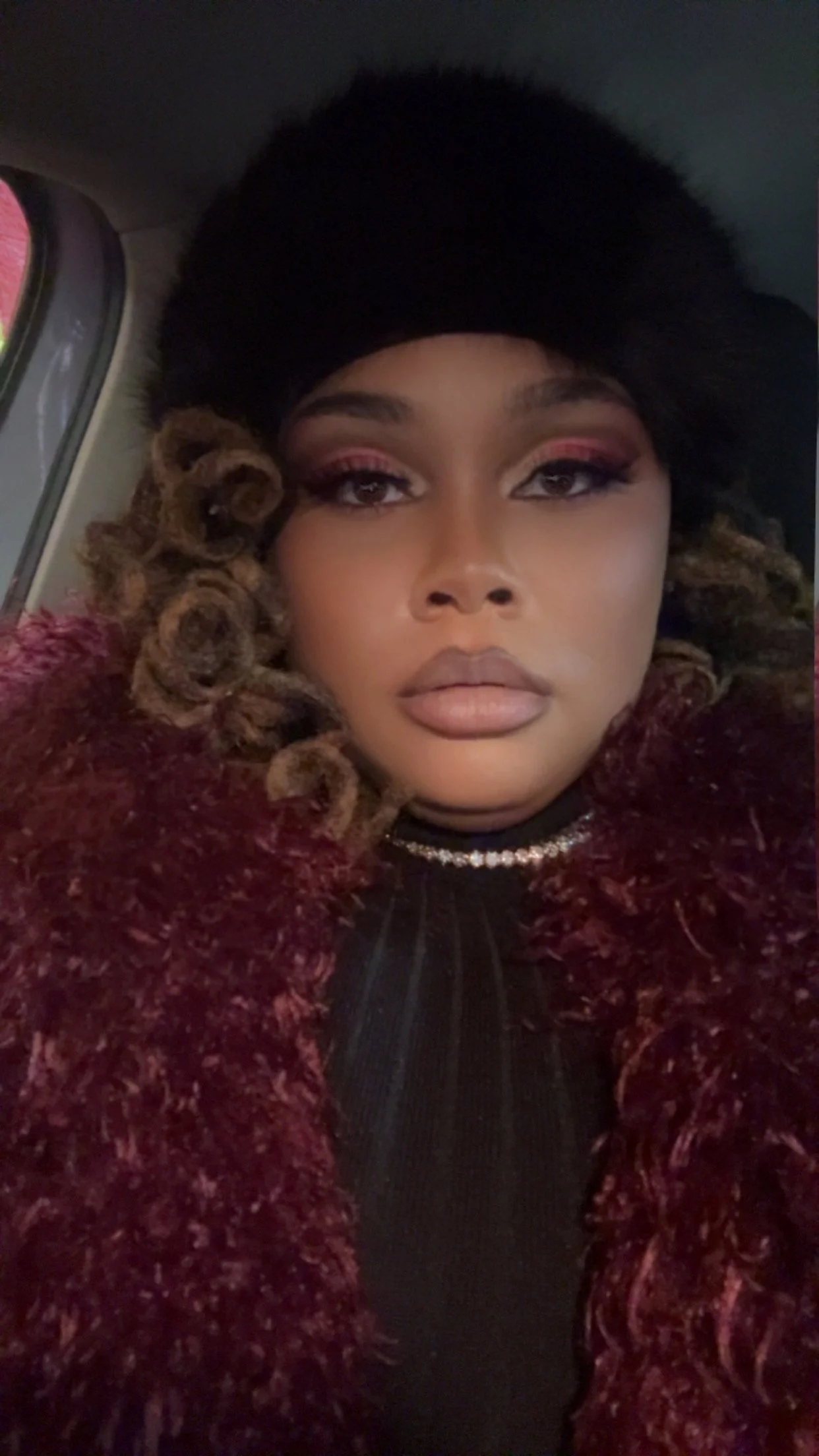 A close-up selfie of a woman with styled hair, wearing a black hat, statement makeup, and a red fur coat, sitting inside a vehicle.