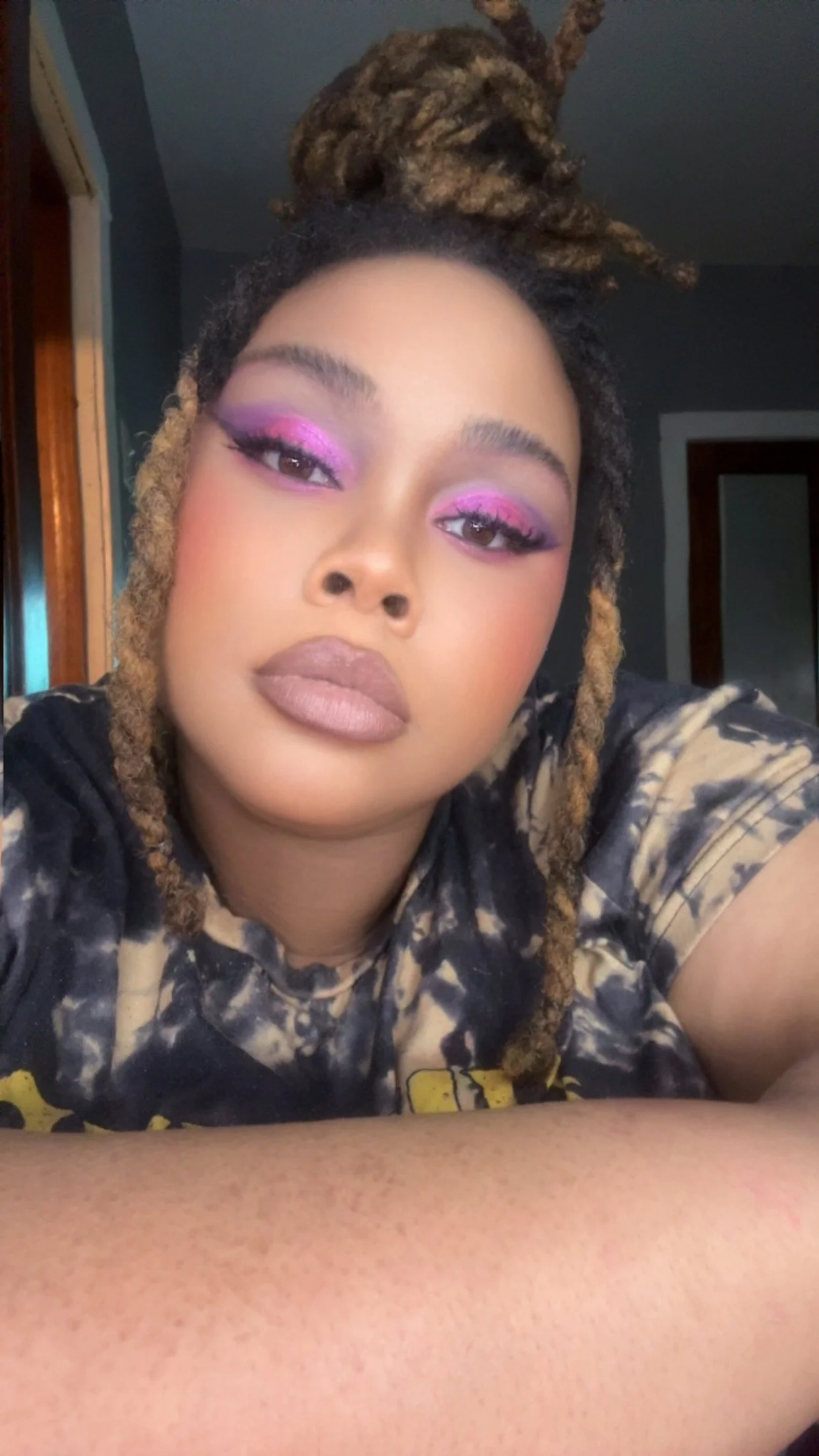 A young woman with makeup, including pink eyeshadow and nude lipstick, and dreadlocks, looks into the camera in a close-up selfie.
