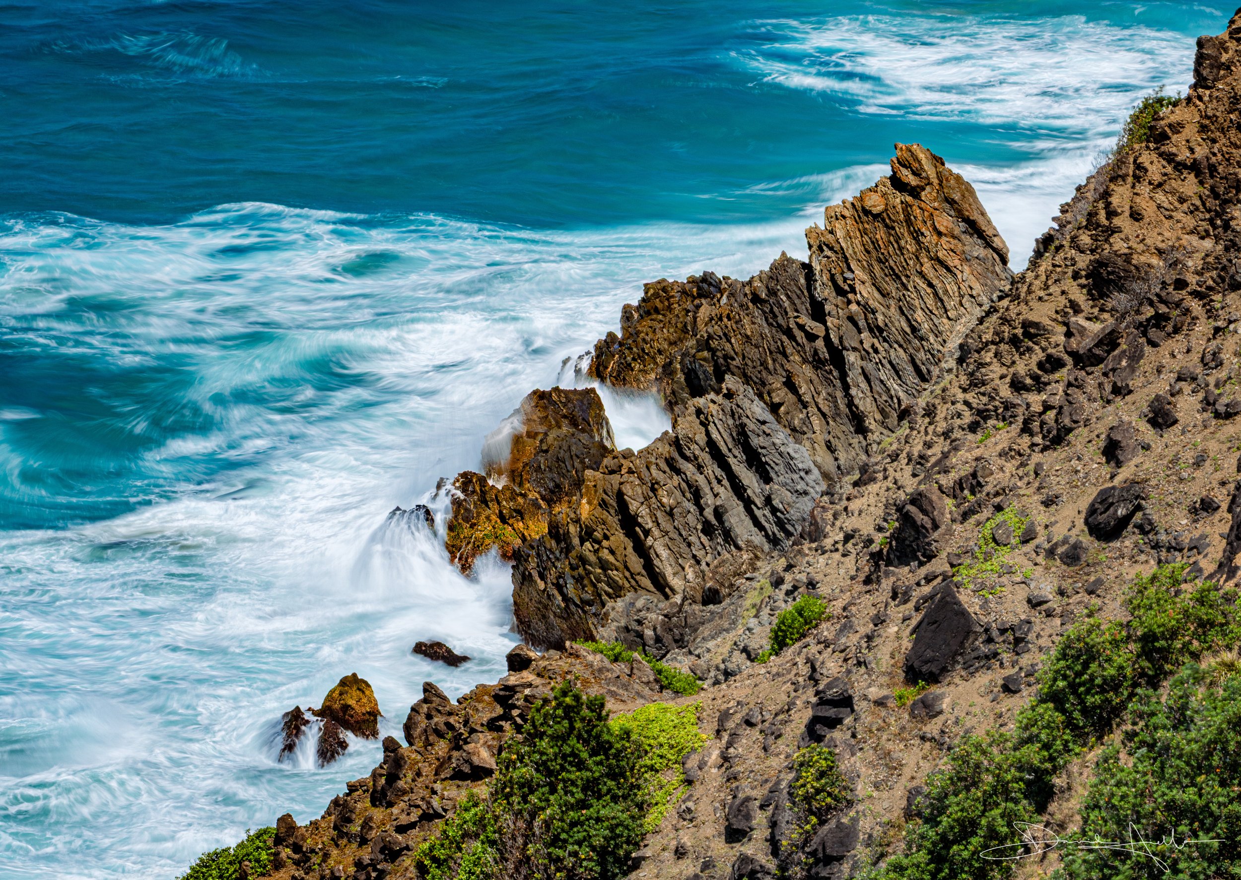A rugged coastal landscape with brown soil and rocks on a cliffside, green bushes at the bottom, and the blue ocean with waves crashing against the rocks.