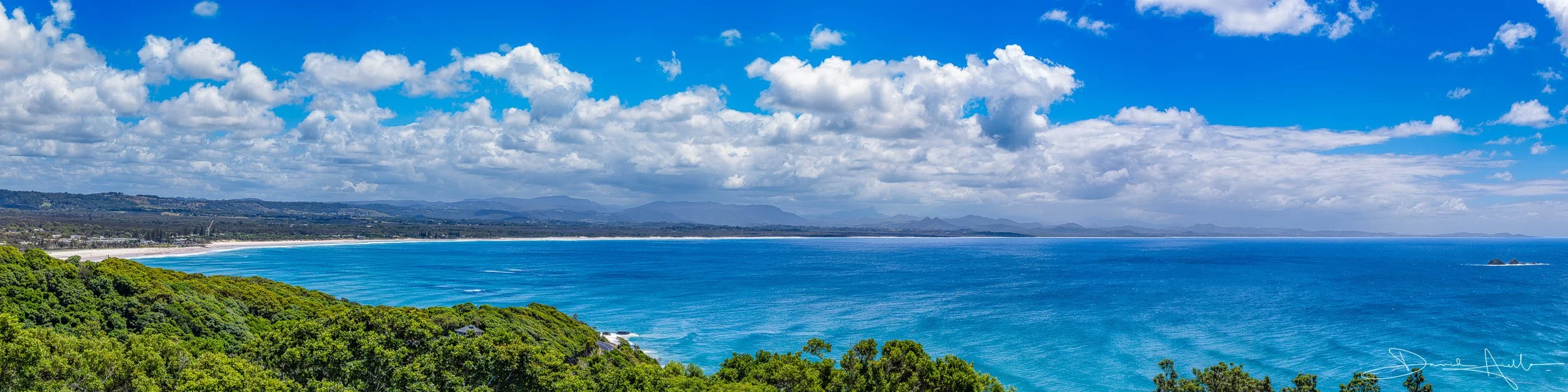 A scenic view of a coastline with lush green trees, blue ocean water, and a partly cloudy sky.