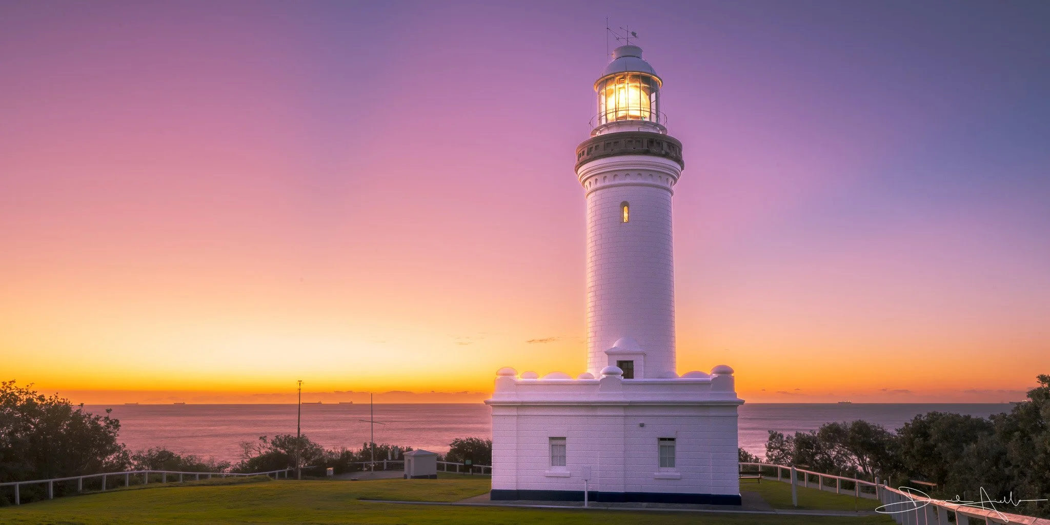 A white lighthouse with a glowing light at sunset, overlooking the ocean with a colorful sky.