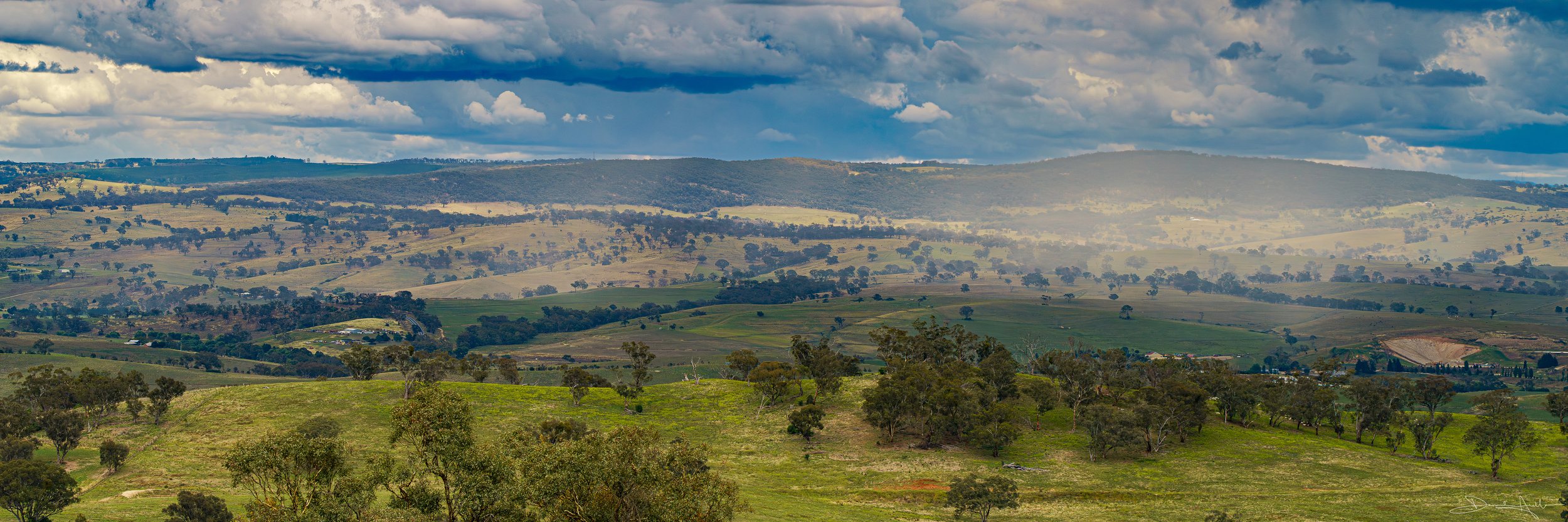 A panoramic view of rolling hills with scattered trees under a partly cloudy sky.