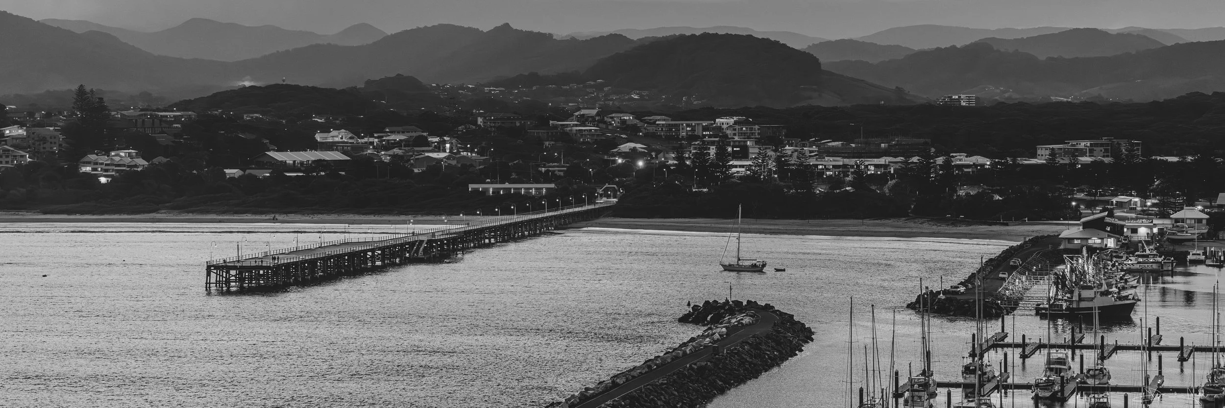 A black and white photo of a coastal town with a pier extending into calm water, boats docked at the marina, residential buildings along the shoreline, and mountains in the background.