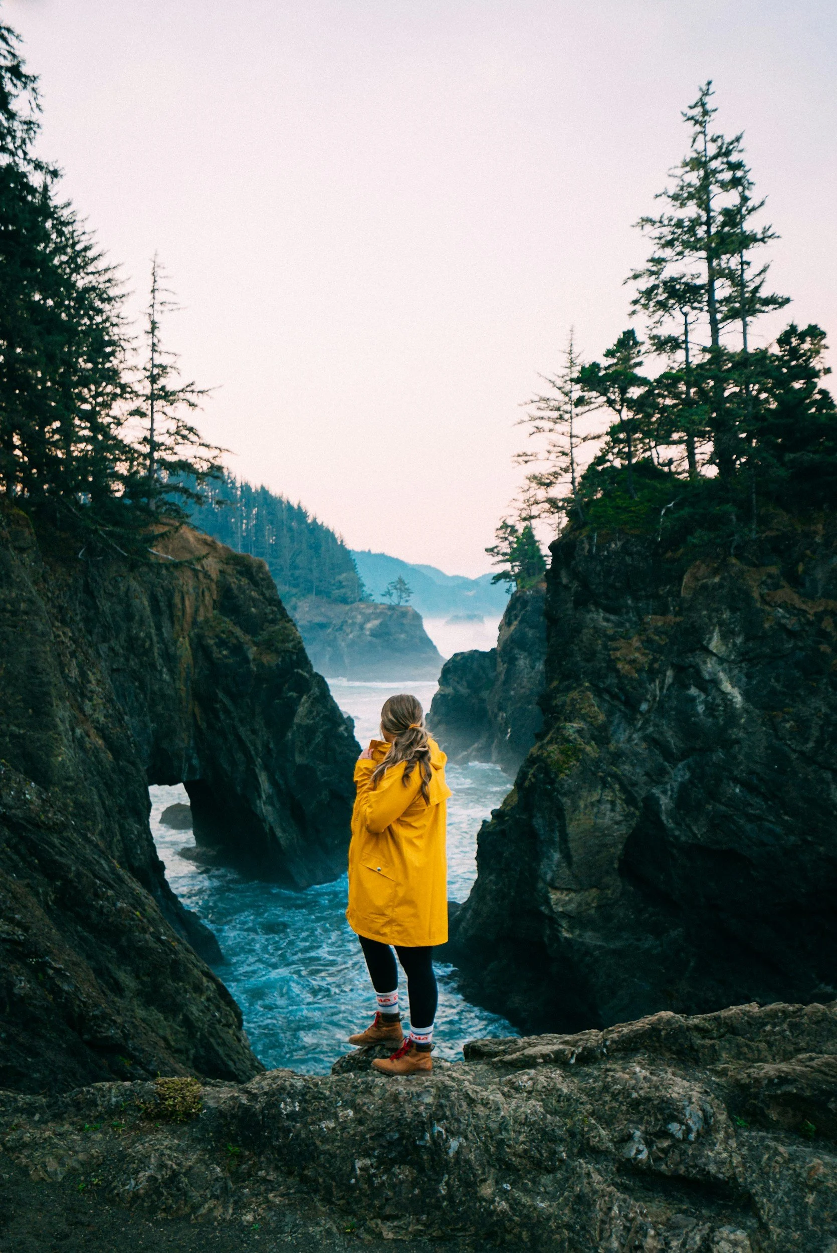 A person in a yellow raincoat standing on rocks between cliffs overlooking the ocean and misty mountains in the distance, with evergreen trees on either side.