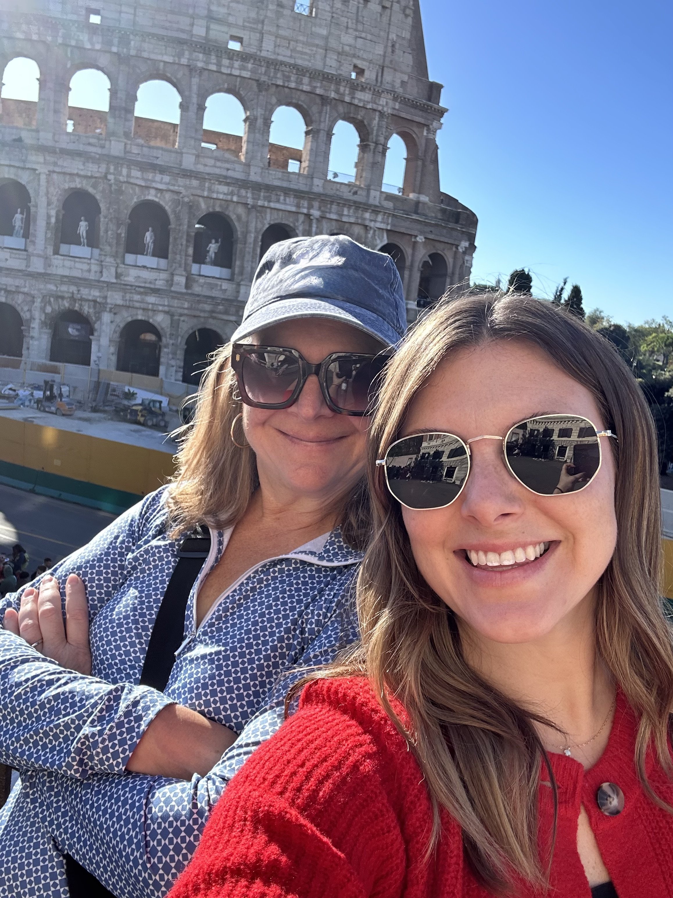 Two women taking a selfie in front of the Colosseum in Rome, Italy. The woman on the left is wearing a blue and white patterned shirt, sunglasses, and a blue hat. The woman on the right is wearing sunglasses and a red sweater. Both are smiling.