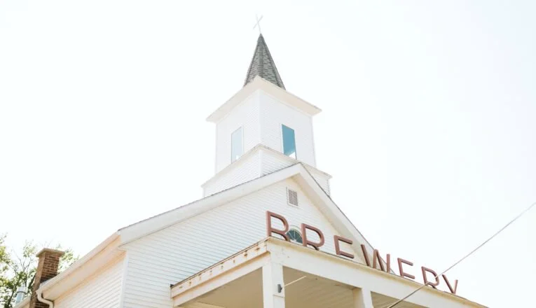 Exterior view of a white church building with a tall steeple and cross on top, with the word "BREWERY" displayed on the front.