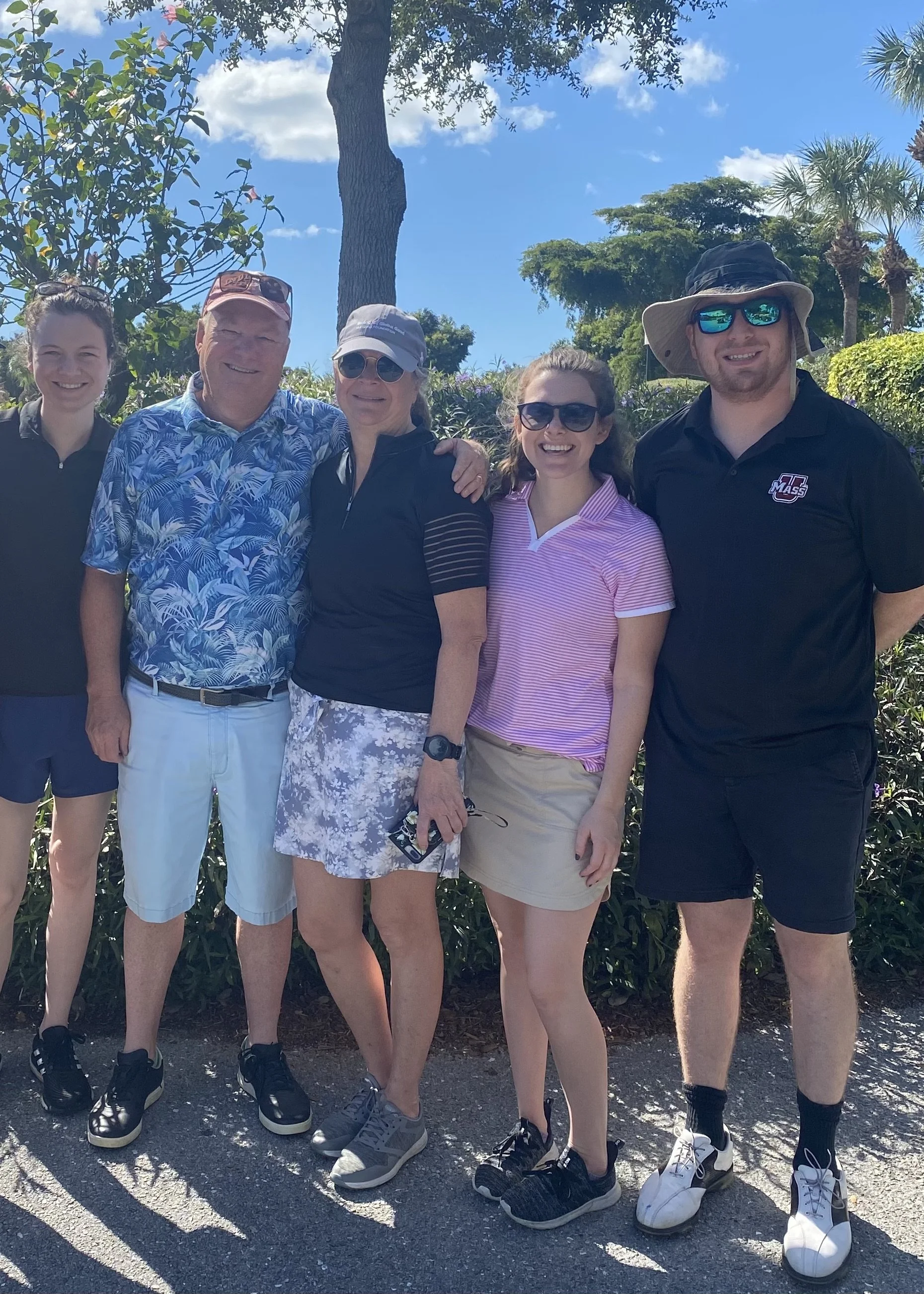Group of six people smiling and standing close together outdoors on a sunny day, with trees and blue sky in the background.