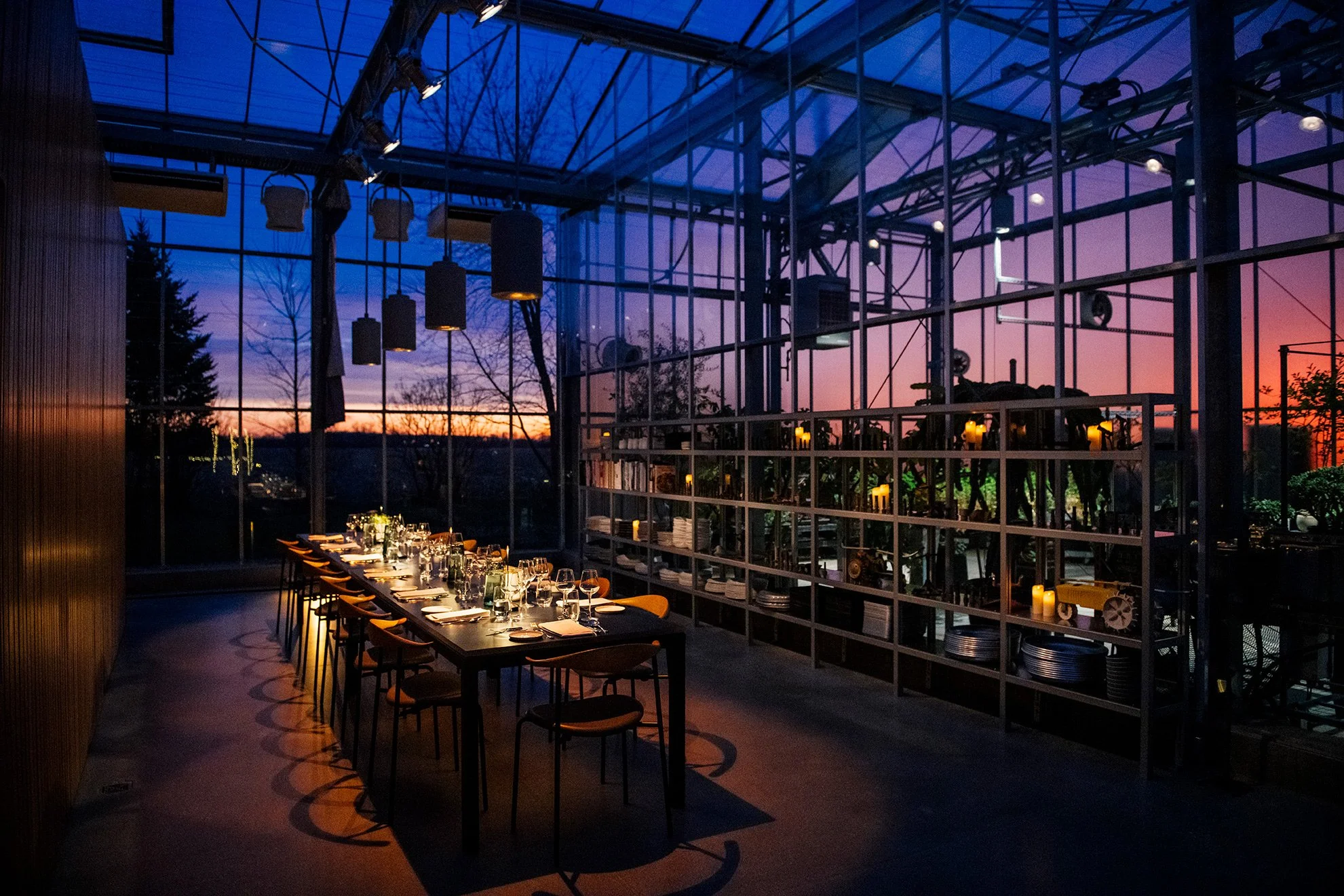 Interior of a modern greenhouse-style restaurant with a long dining table set for a meal, with glasses, plates, and cutlery, overlooking a sunset sky through large glass windows.