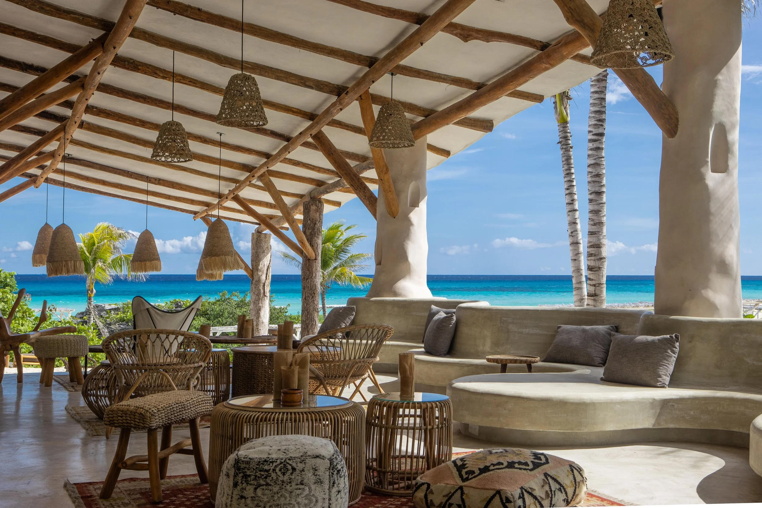 A beachside resort lounge with woven hanging lights, wicker chairs, and a cushioned seating area, overlooking the ocean with palm trees under a clear blue sky- beautiful location for a Mahjong Retreat!