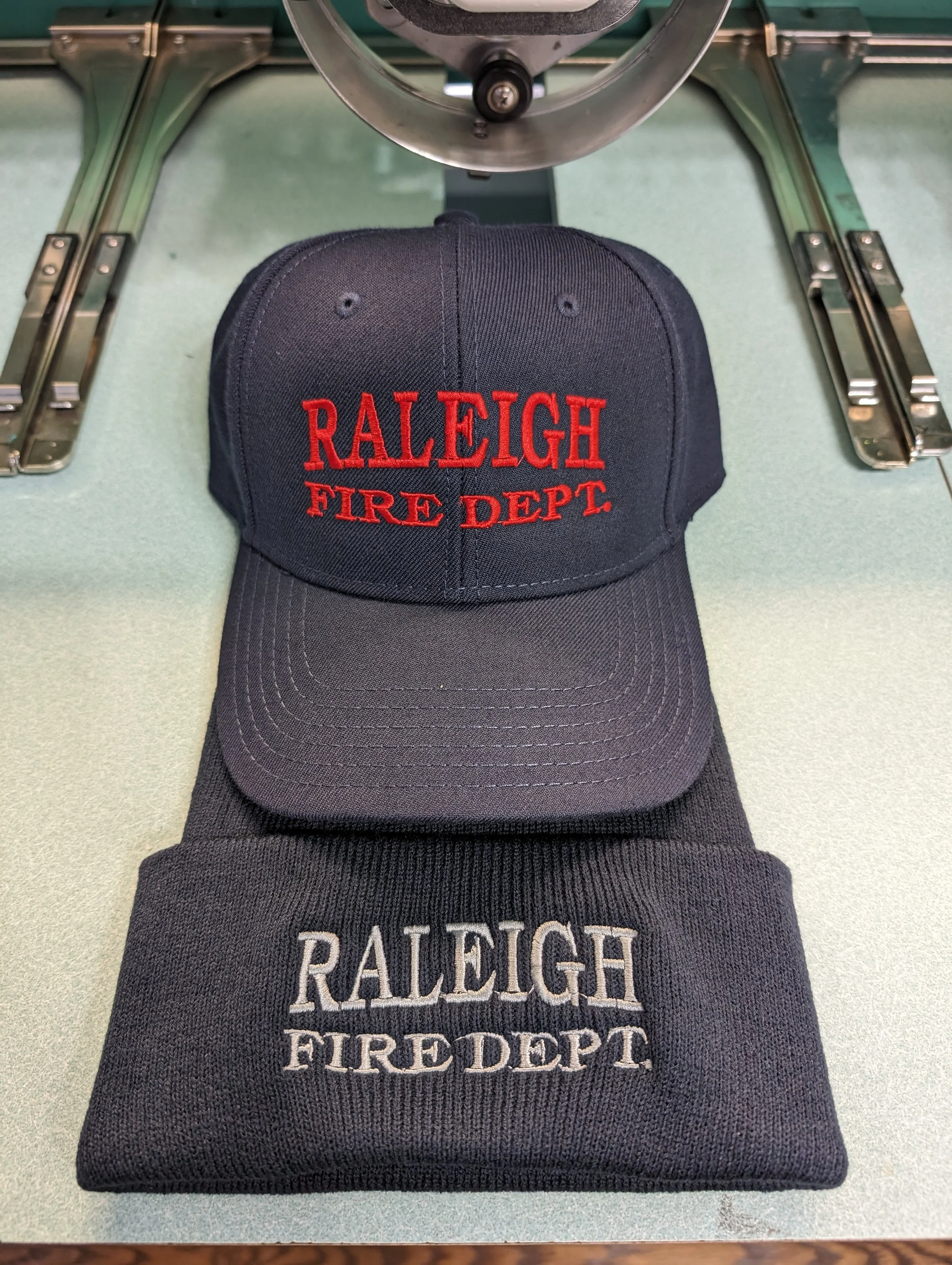 A black baseball cap and a black knit beanie, both embroidered with 'Raleigh Fire Dept' in red and white thread respectively, placed on a green surface with a metal object above.