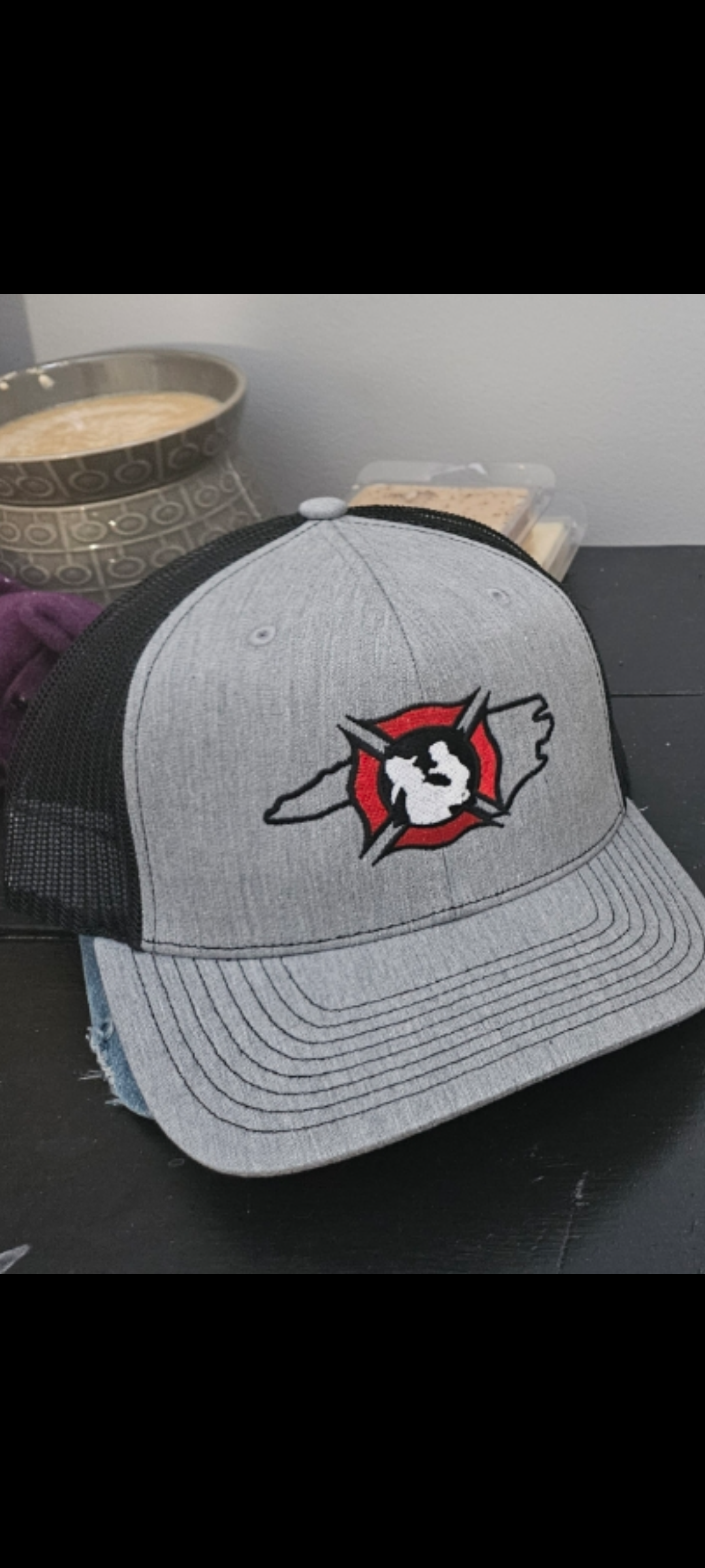 Gray and black baseball cap with a logo of a snarling wolf's head inside a red circle with lightning bolts on a black surface, with a cup of coffee and a container in the background.