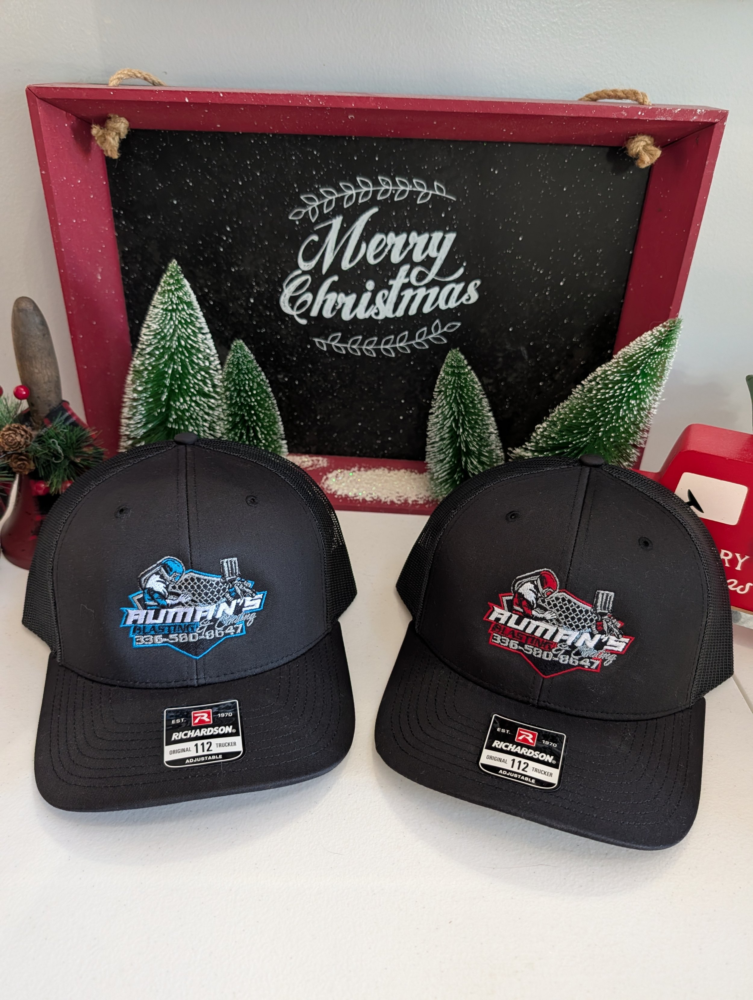 Two baseball caps with logos, one blue and white, the other red and black, placed on a white surface. Behind them, a Christmas decoration with a sign that says 'Merry Christmas' and small snow-covered Christmas trees.