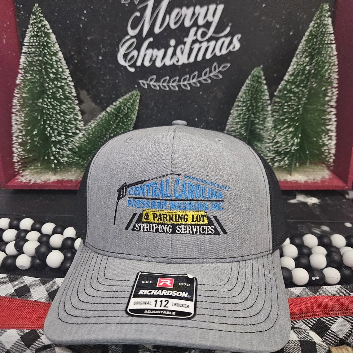 A gray Richardson trucker cap with embroidery of Central Carolina Pressure Washing Inc., and a yellow patch for parking lot striping services. Behind the cap is a Christmas-themed sign with the message "Merry Christmas" and snow-dusted green pine tre