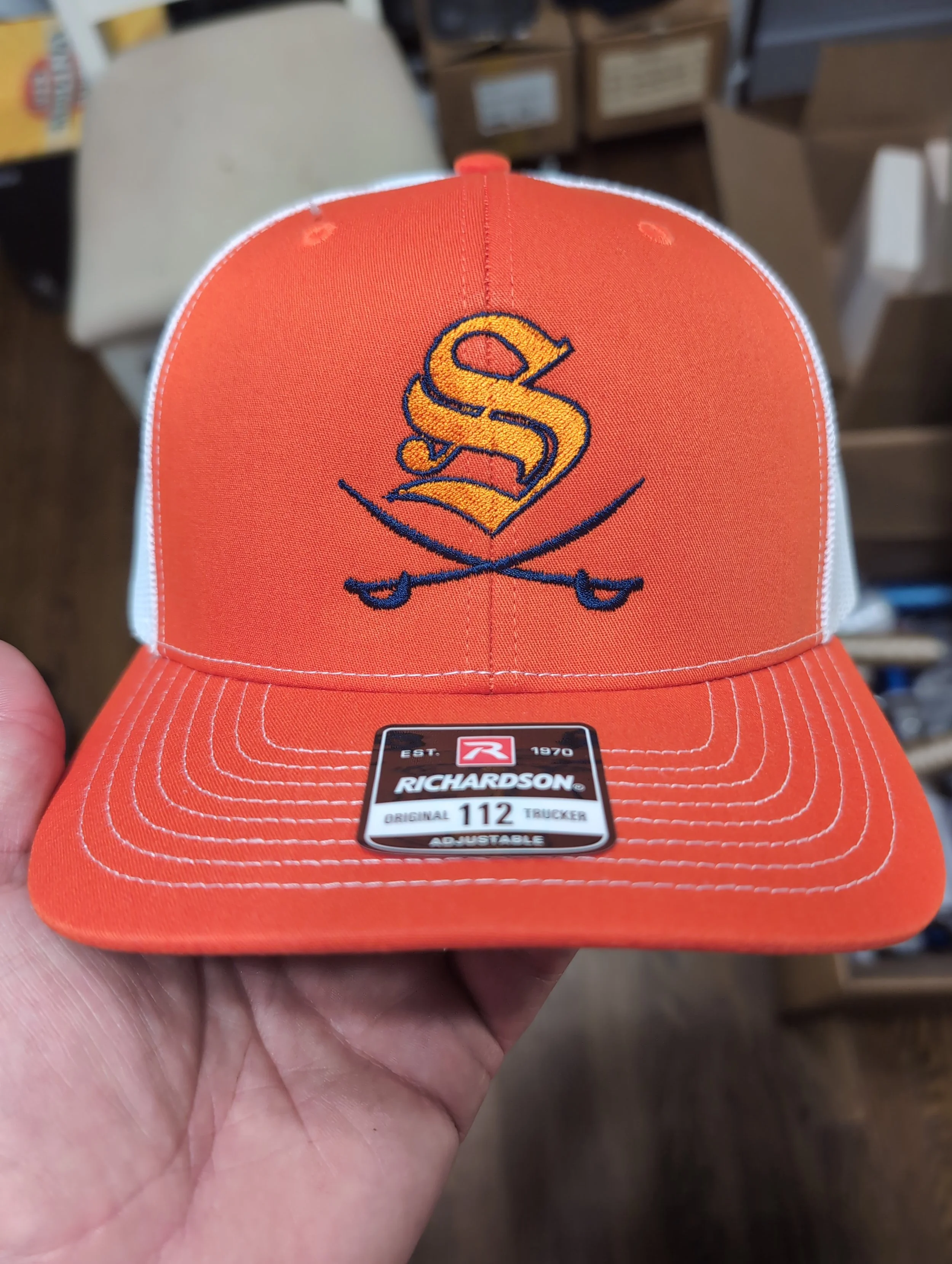 Orange trucker hat with embroidered logo of a yellow S symbol and crossed golf clubs, and white stitching on the brim.