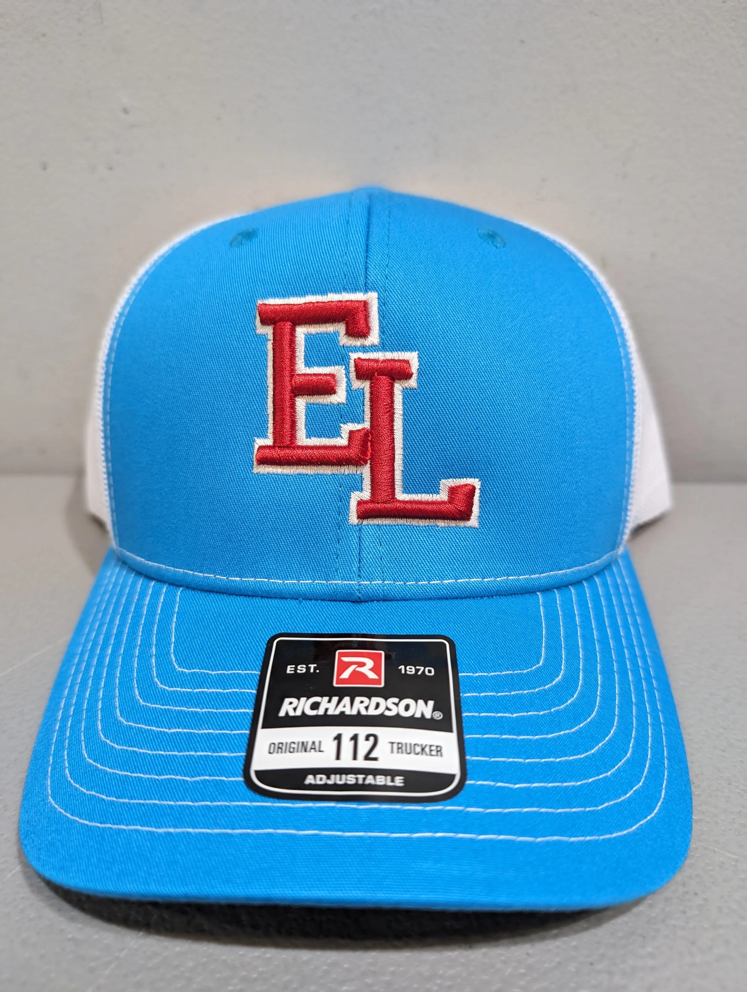 A blue and white Richardson trucker hat with red embroidered initials 'EL' on the front and a black sticker on the brim that says 'Richardson, established 1970, adjustable, original 112 trucker.'