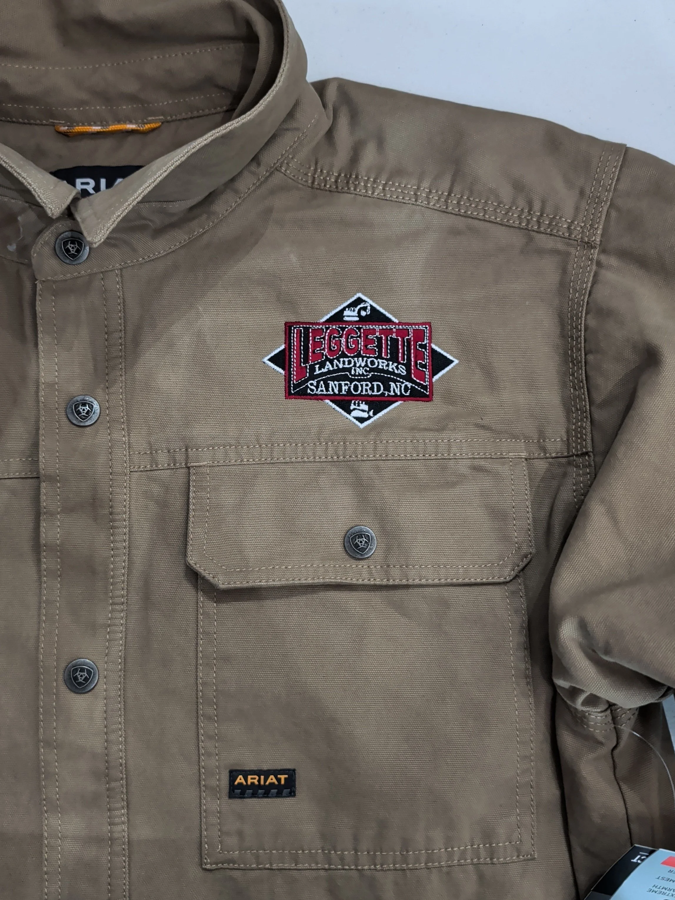 Close-up of a tan work jacket with an embroidered patch reading "Leggett Landworks Inc. Sanford, NC" on the chest pocket, and an orange and black "Ariat" label on the pocket.