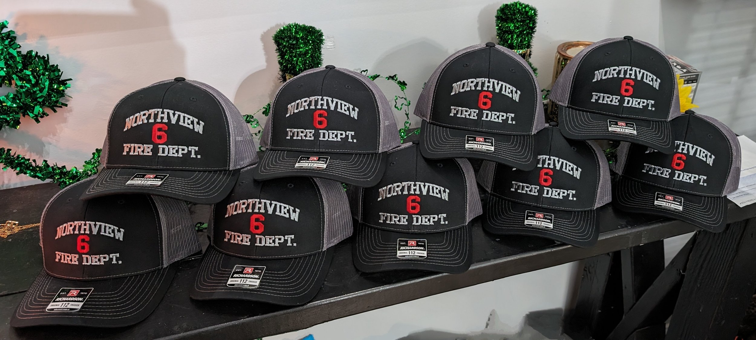 A collection of black and gray trucker caps with embroidery that reads "NORTHVIEW 6 FIRE DEPT," arranged on a black table with holiday decorations in the background.