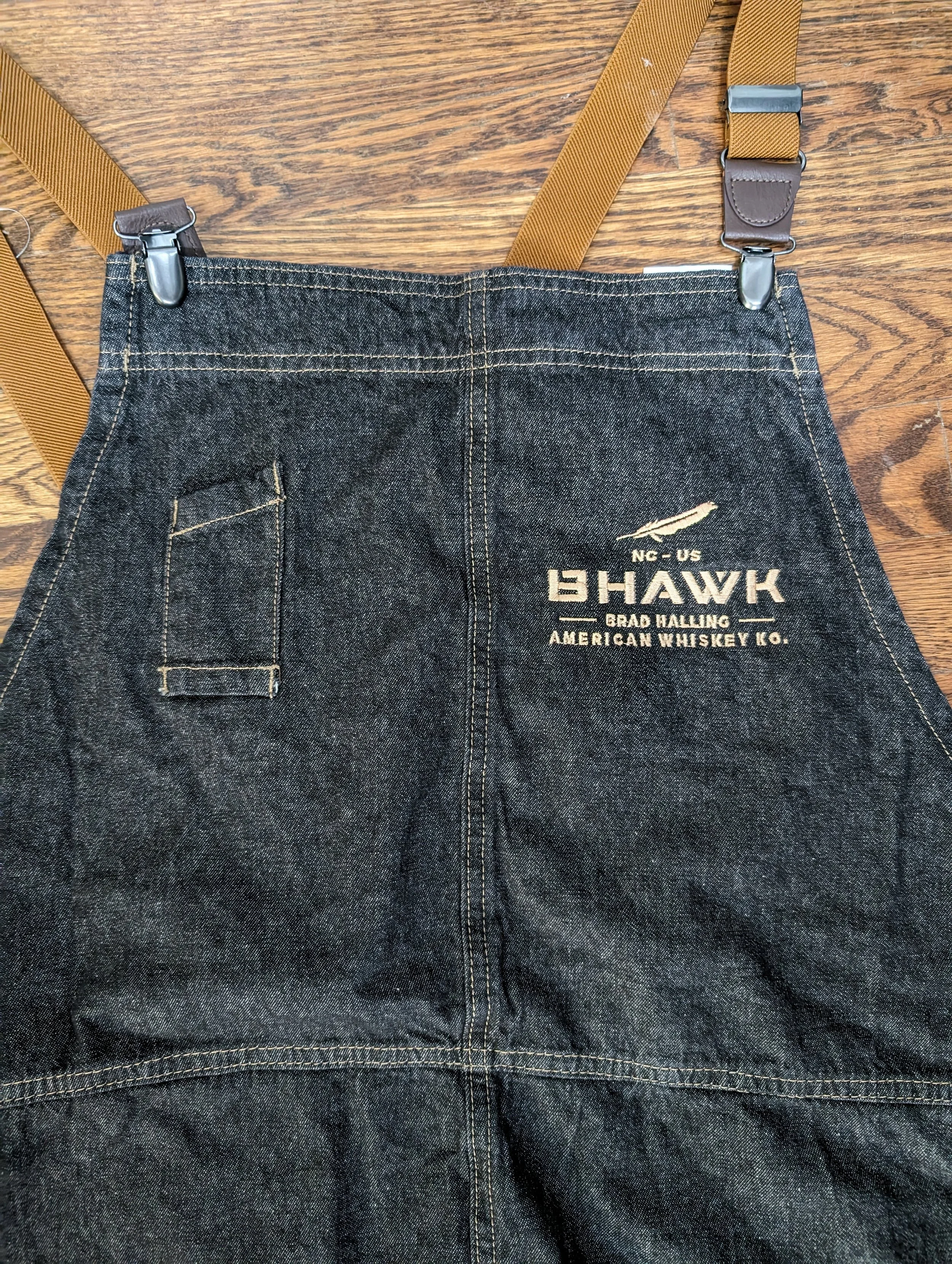 Black denim apron with a company logo and text, featuring adjustable tan straps, laid on a wooden surface.