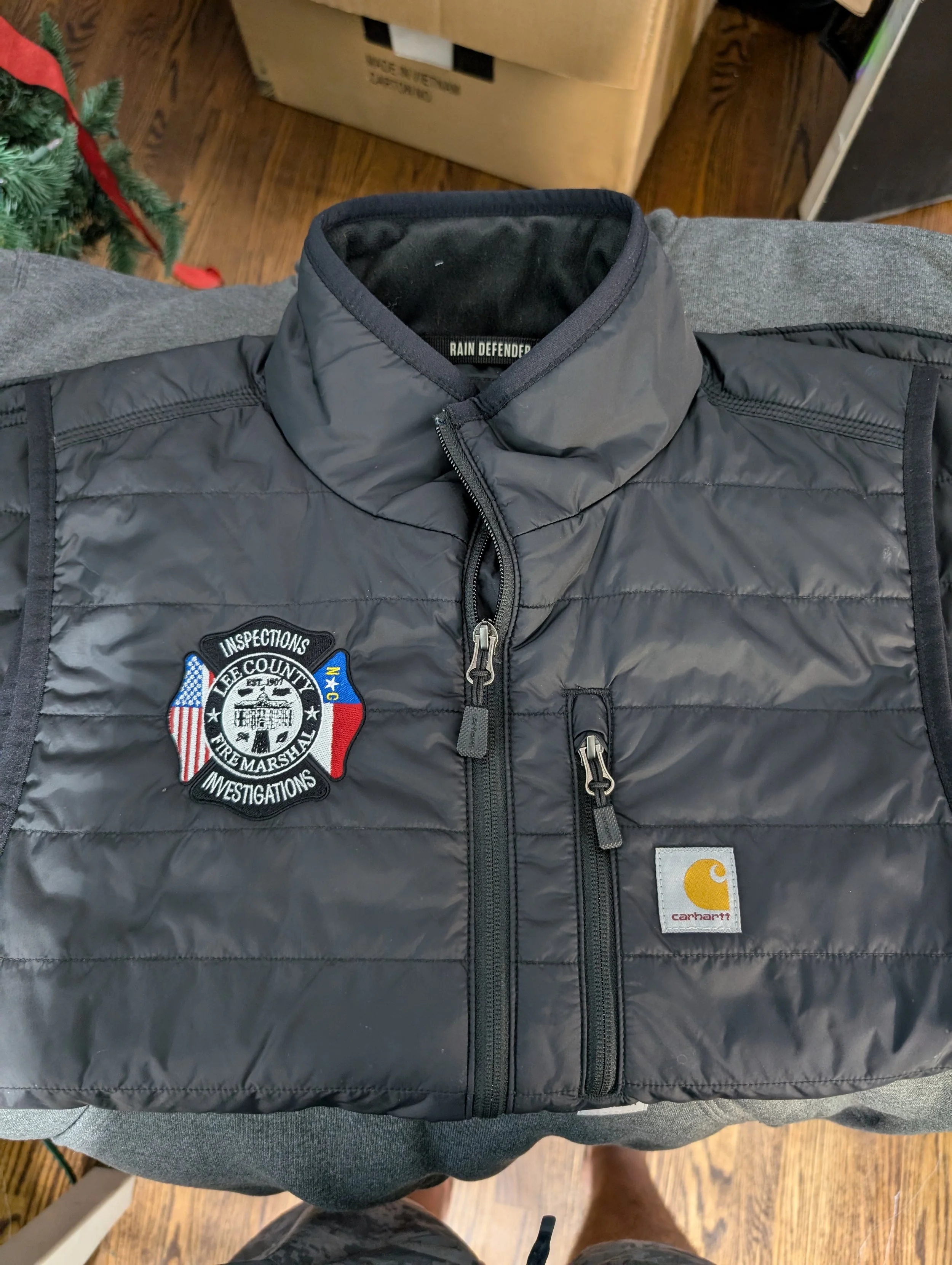 A gray Carhartt jacket with police badges and patches, including one for Lee County Fire Marshal Inspections, lying on a person's lap.